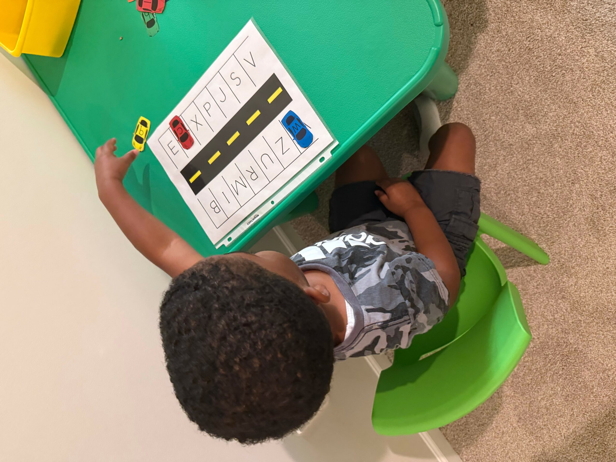Child sitting at a green table with a whiteboard showing a road sign and traffic lights, holding a card.