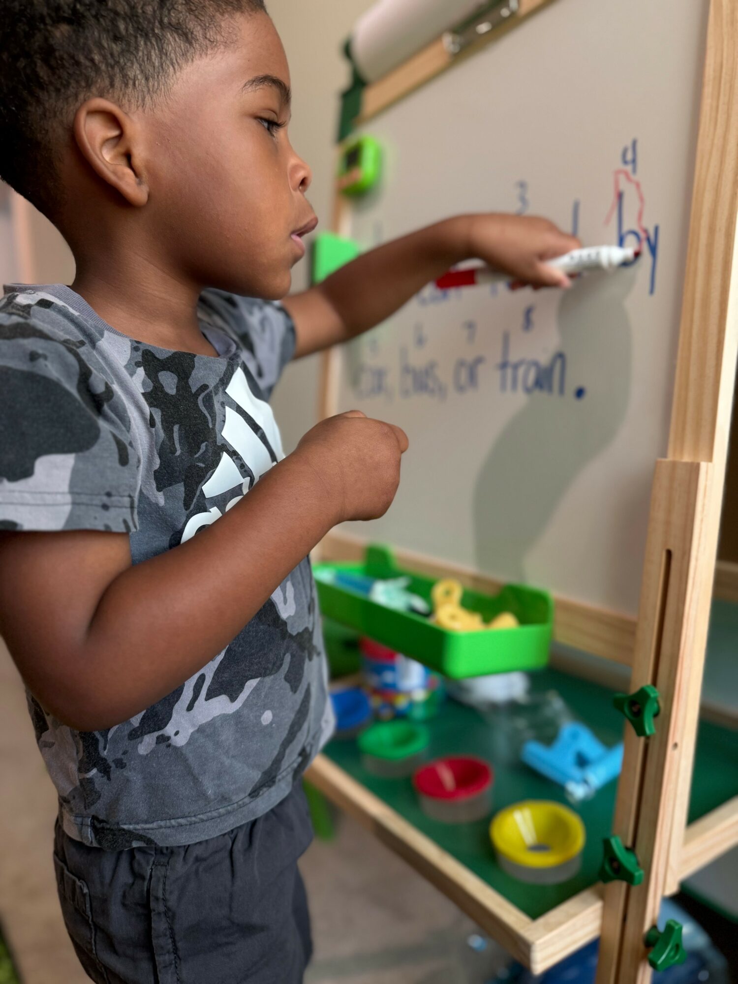 Child writing on a whiteboard with markers, standing near a green tray of colorful toys and tools, in a classroom.