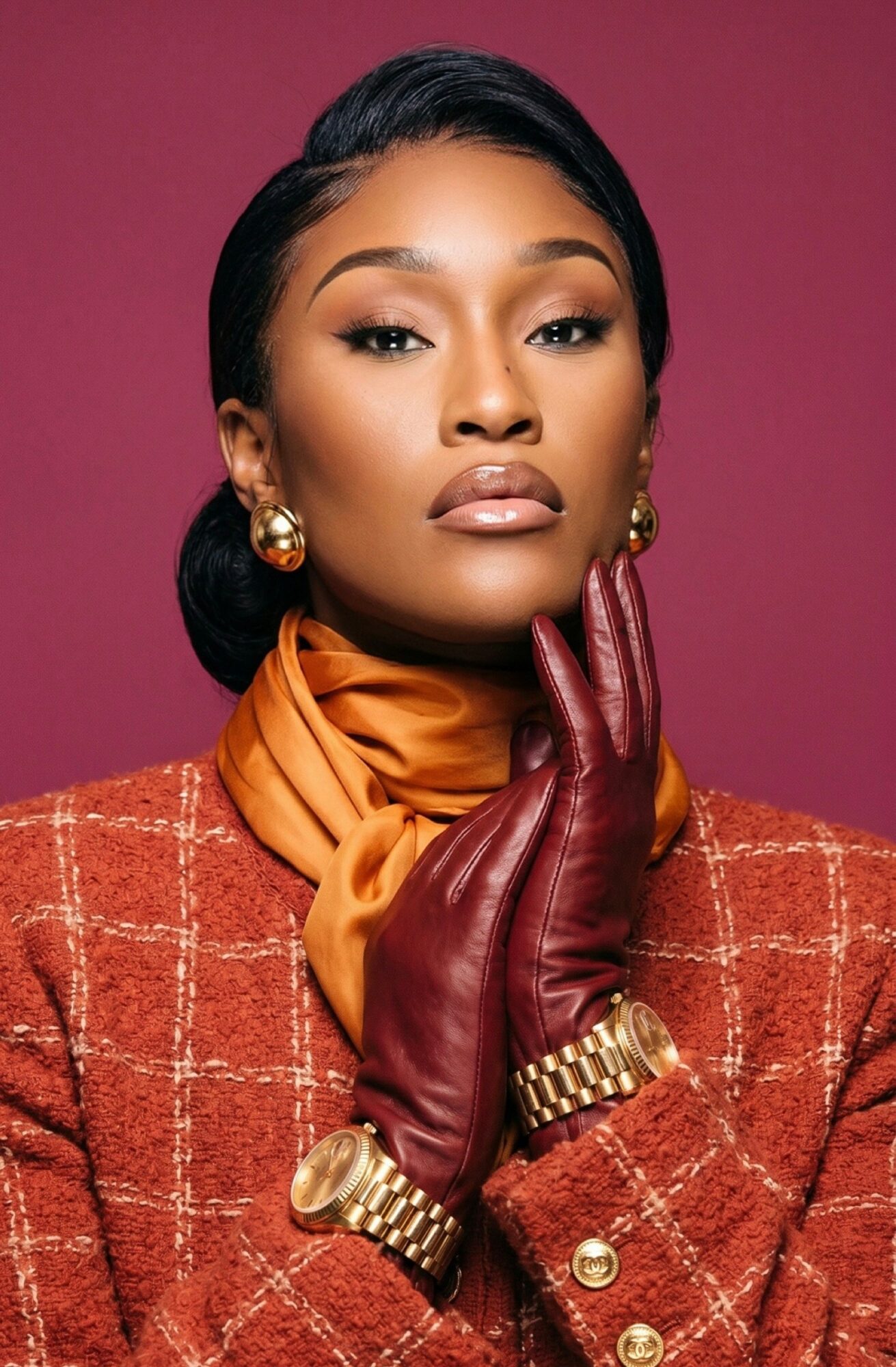 Woman with styled hair, wearing a checkered blazer, gold jewelry, orange scarf, and red gloves, posing against a pink background.