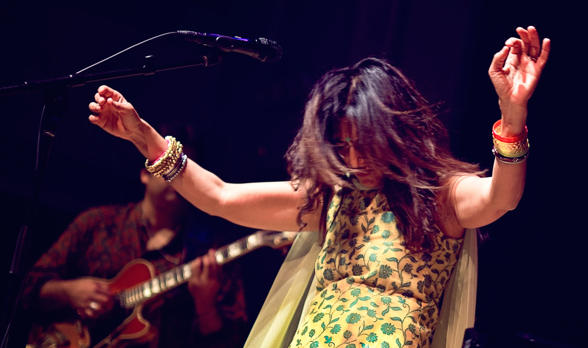 Woman with arms raised dancing on stage, musician playing guitar in background, dark stage setting