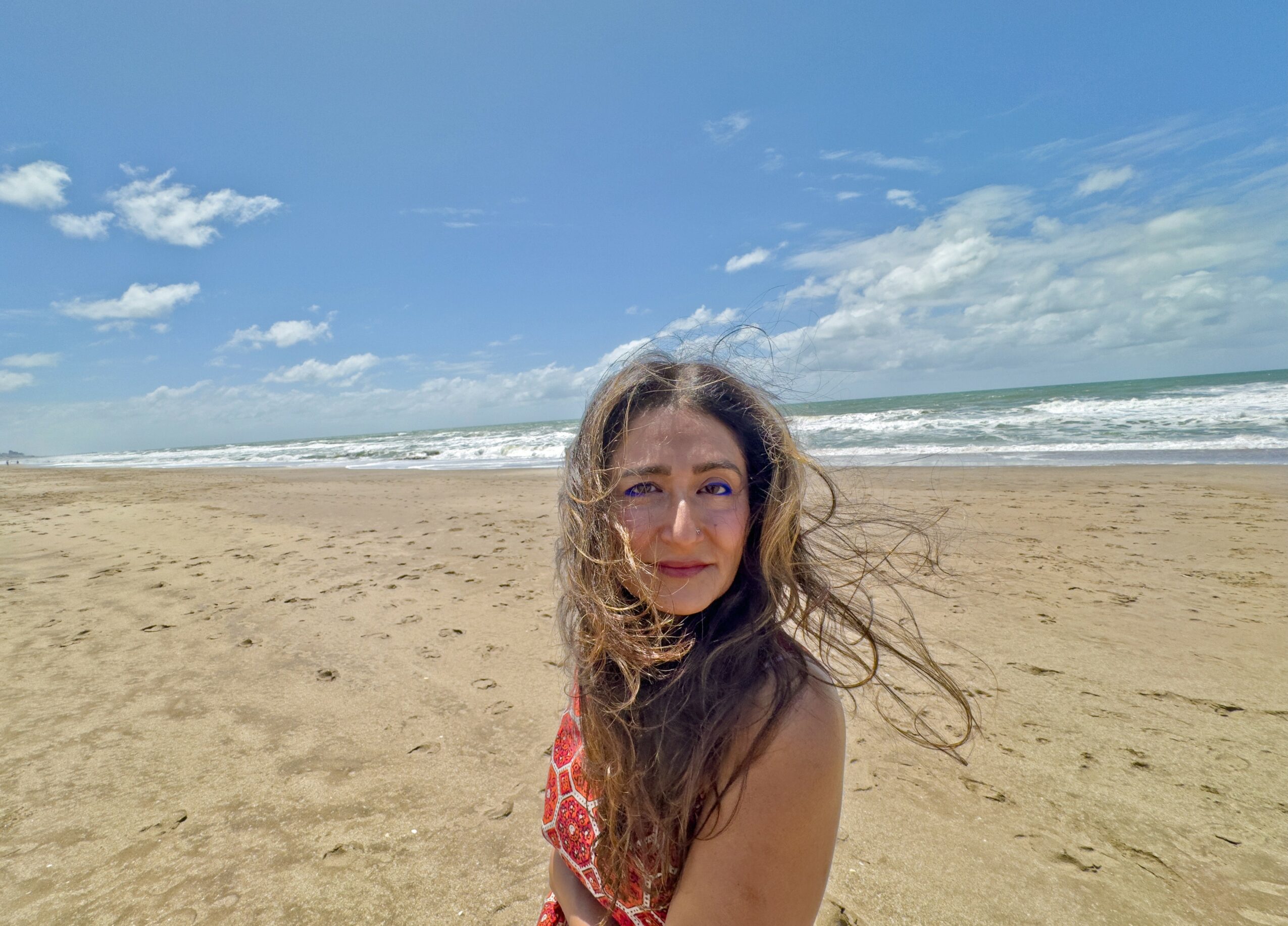 Young woman with curly hair on a sandy beach, ocean and blue sky in background.