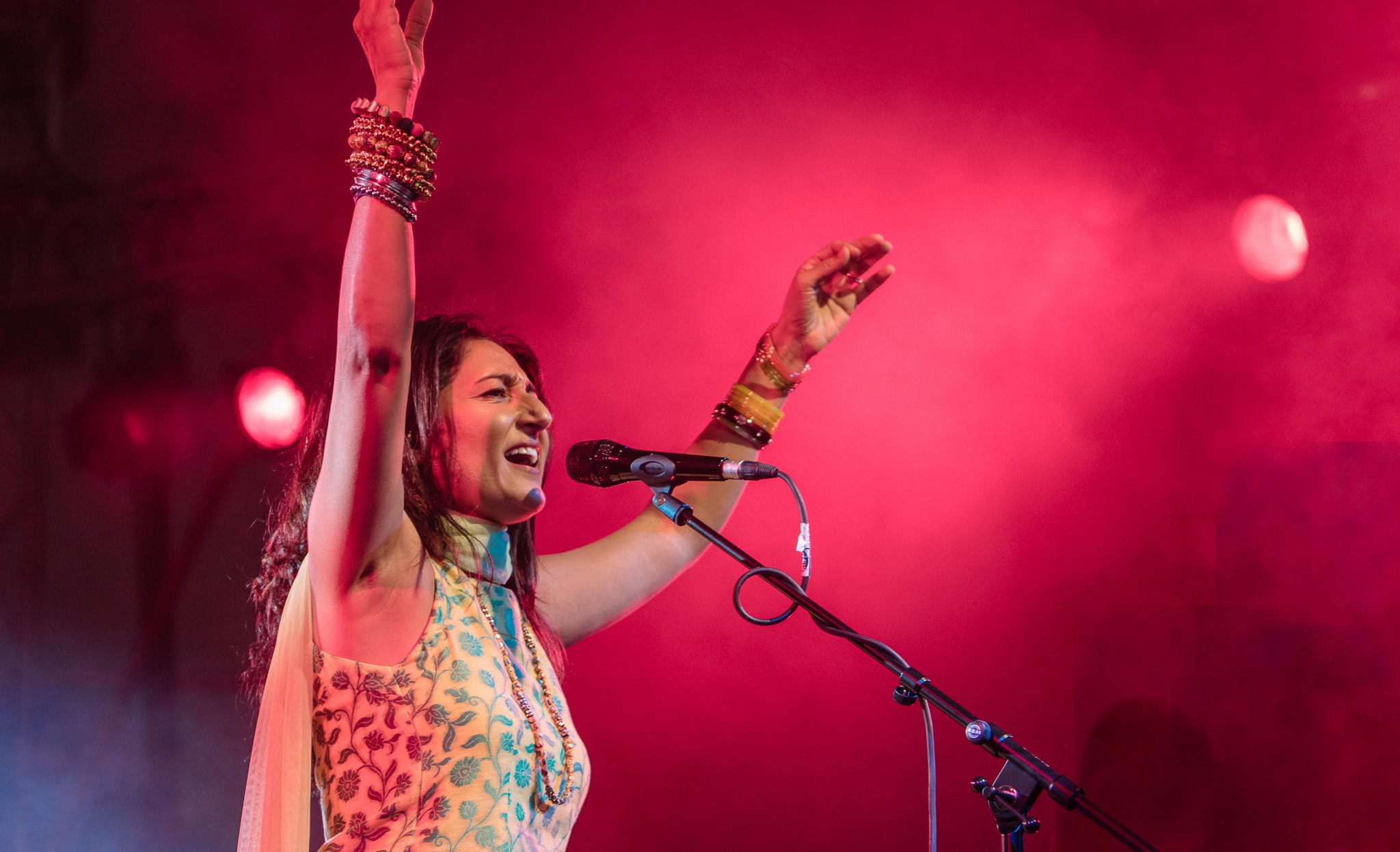 A woman singing into a microphone on stage with pink lighting, raising her arms, wearing colorful clothing and jewelry.