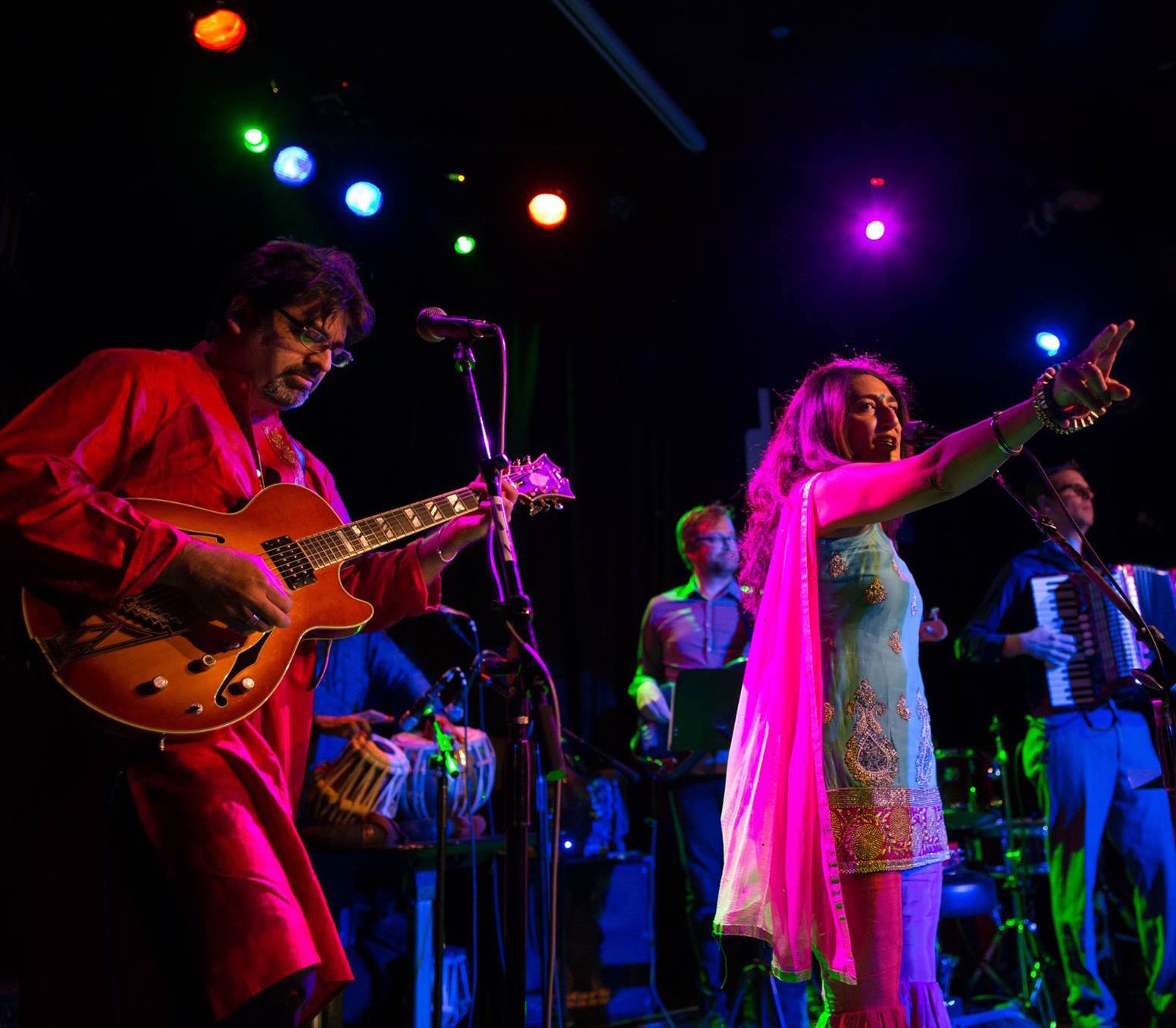 Group of musicians performing on stage with colorful lights, including a woman singing and a man playing guitar.