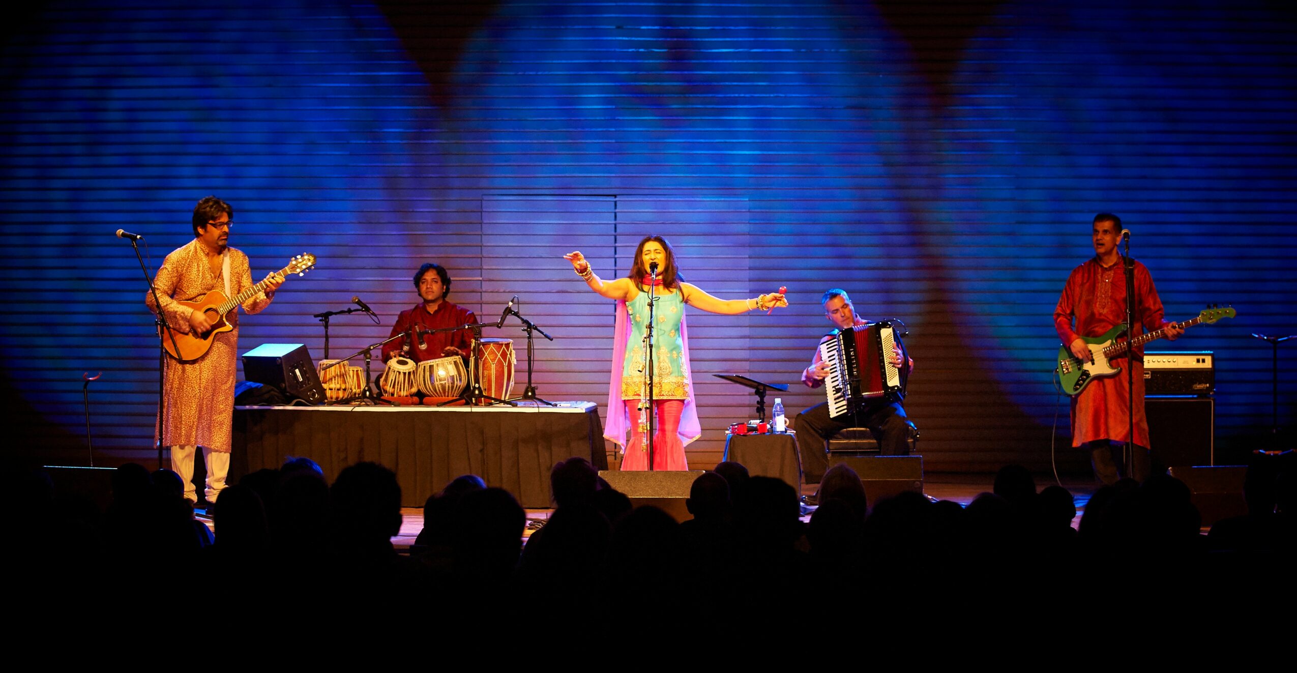 Musicians perform on stage with a singer in the center, colorful background, audience in foreground.