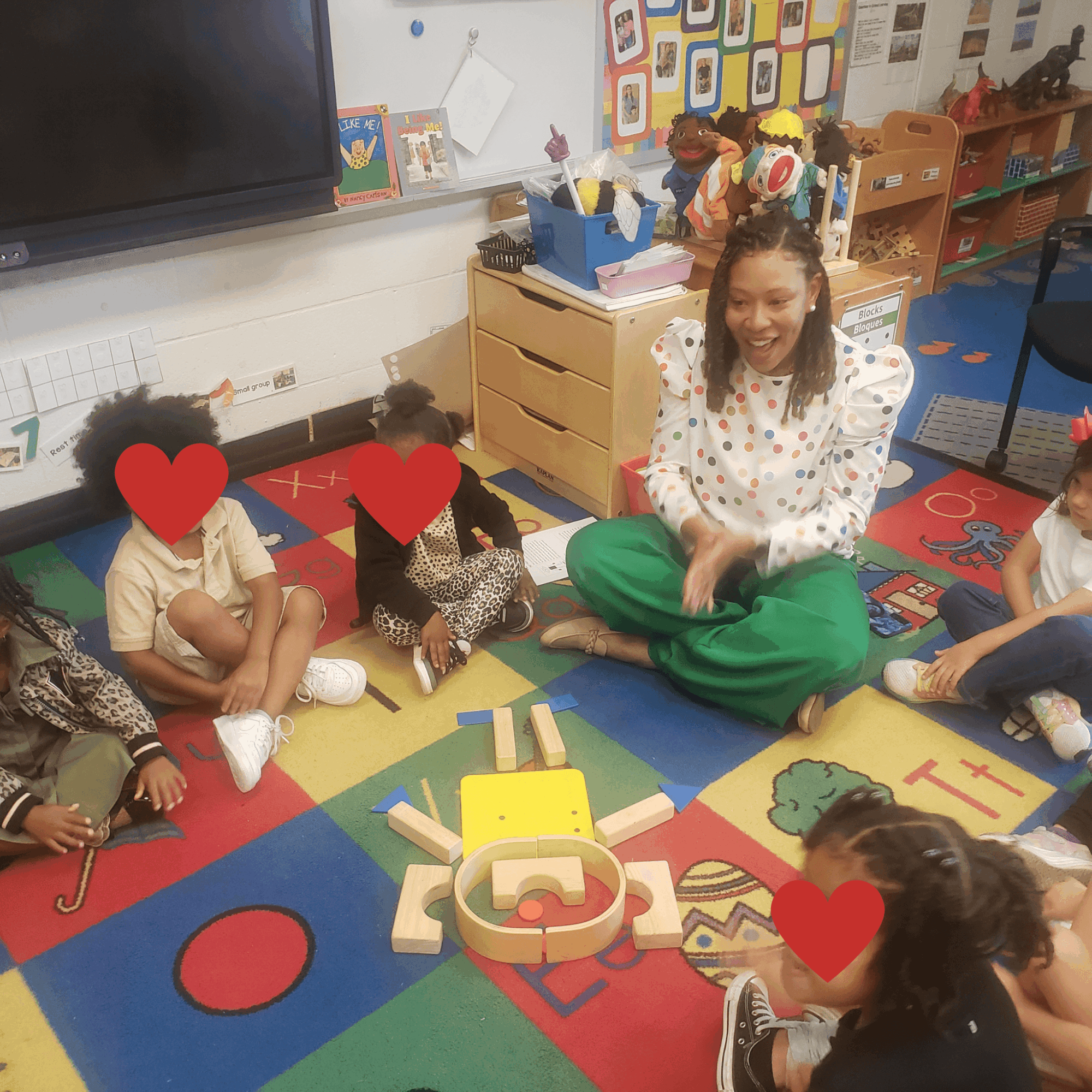 A teacher sitting on a colorful classroom carpet with children, some with faces covered, engaging in an activity.