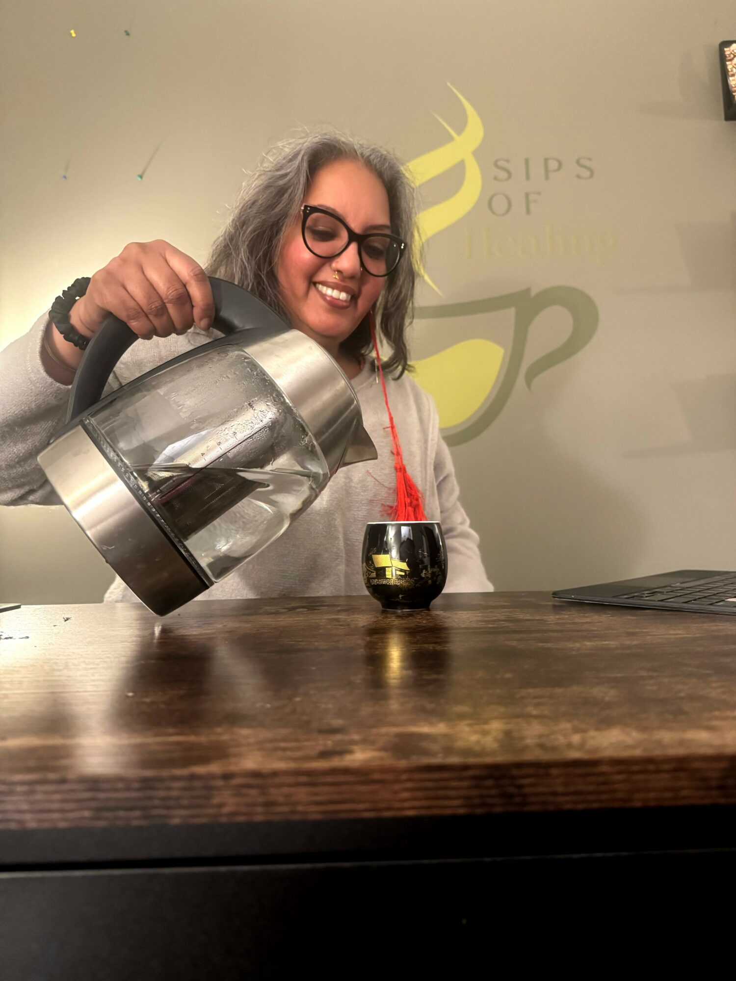 Person with glasses pouring tea from a kettle into a cup on a wooden table, smiling, in a cafe setting.