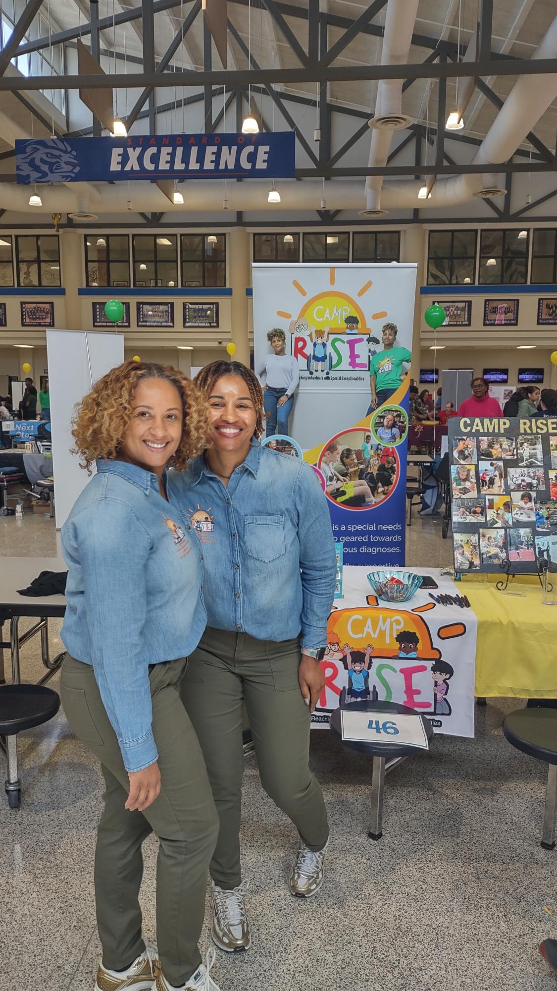 Two smiling women in denim jackets standing in front of a camp booth with colorful posters and a sign that says 'Camp Rise'.