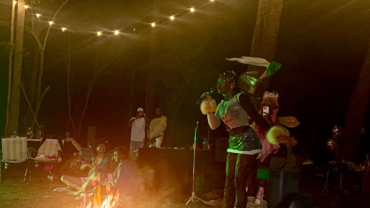 Person in costume holding a ball, with people and tables in a dark outdoor setting, illuminated by string lights.
