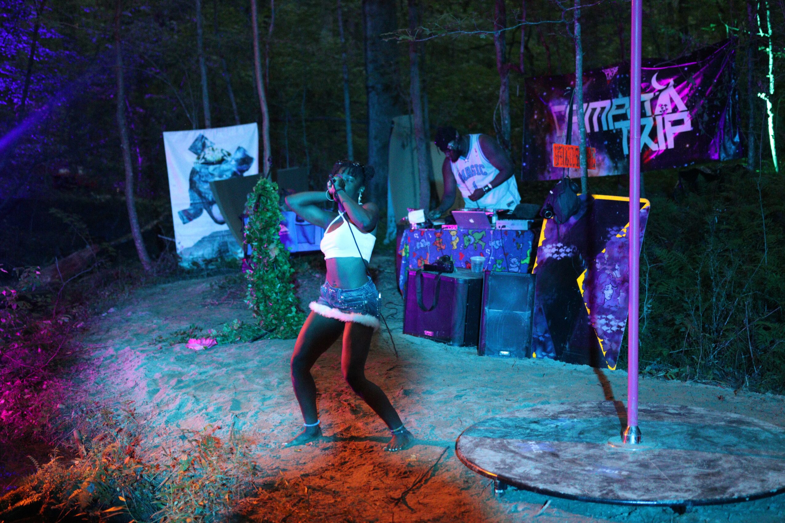 Person dancing outdoors at night with colorful lights and banners, surrounded by trees and equipment, illuminated by vibrant lighting.