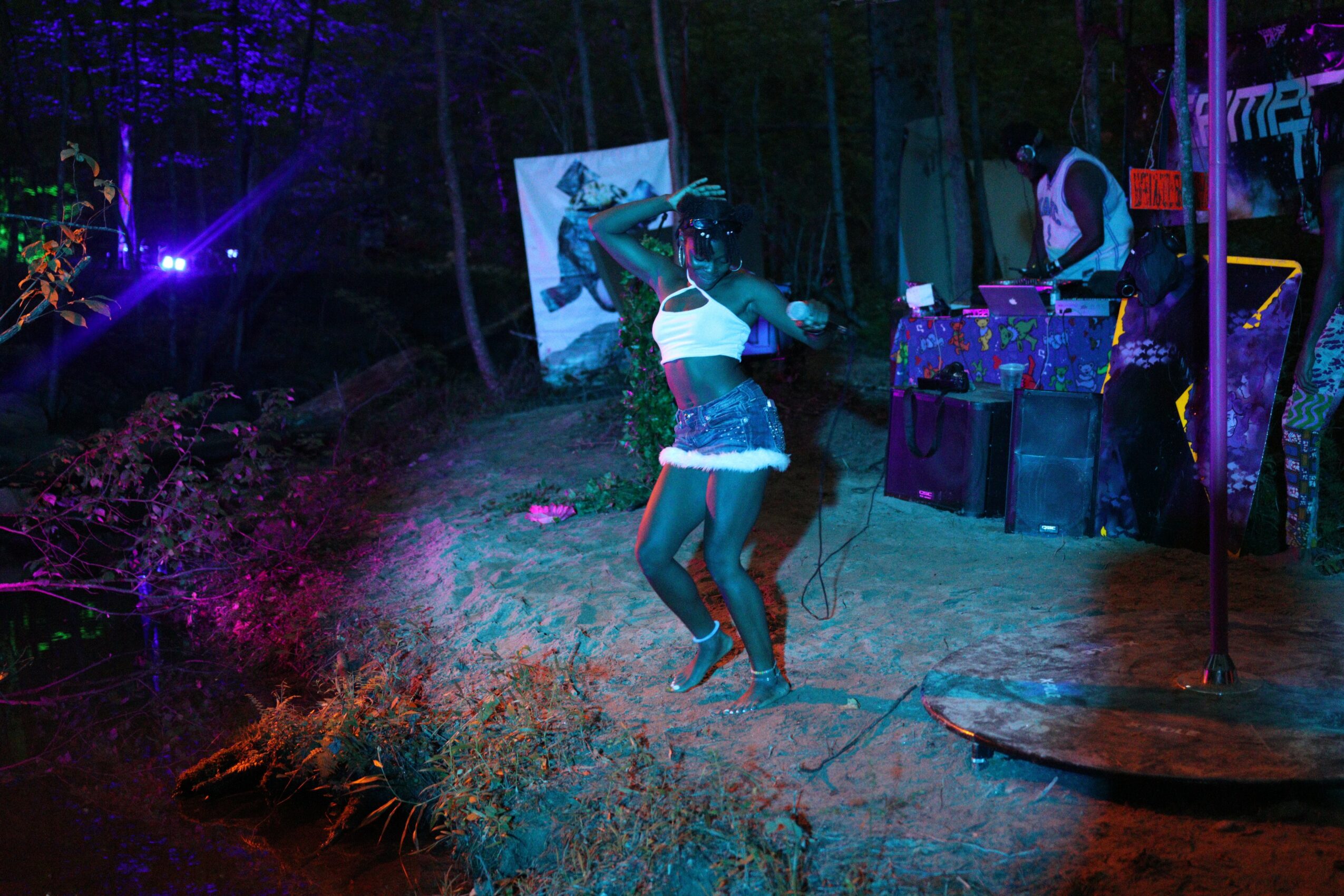 Person dancing outdoors at night with DJ setup and colorful lights, surrounded by trees and equipment.