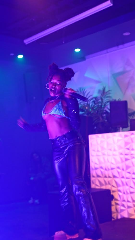 Person dancing on stage with colorful lighting, wearing a shiny outfit and glasses, smiling happily.