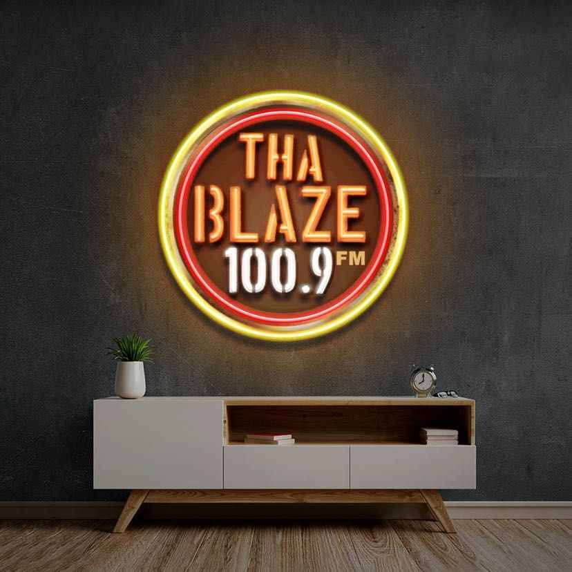 Neon sign reads 'THA BLAZE 100.9 FM' on a dark wall above a white sideboard with a plant and books.