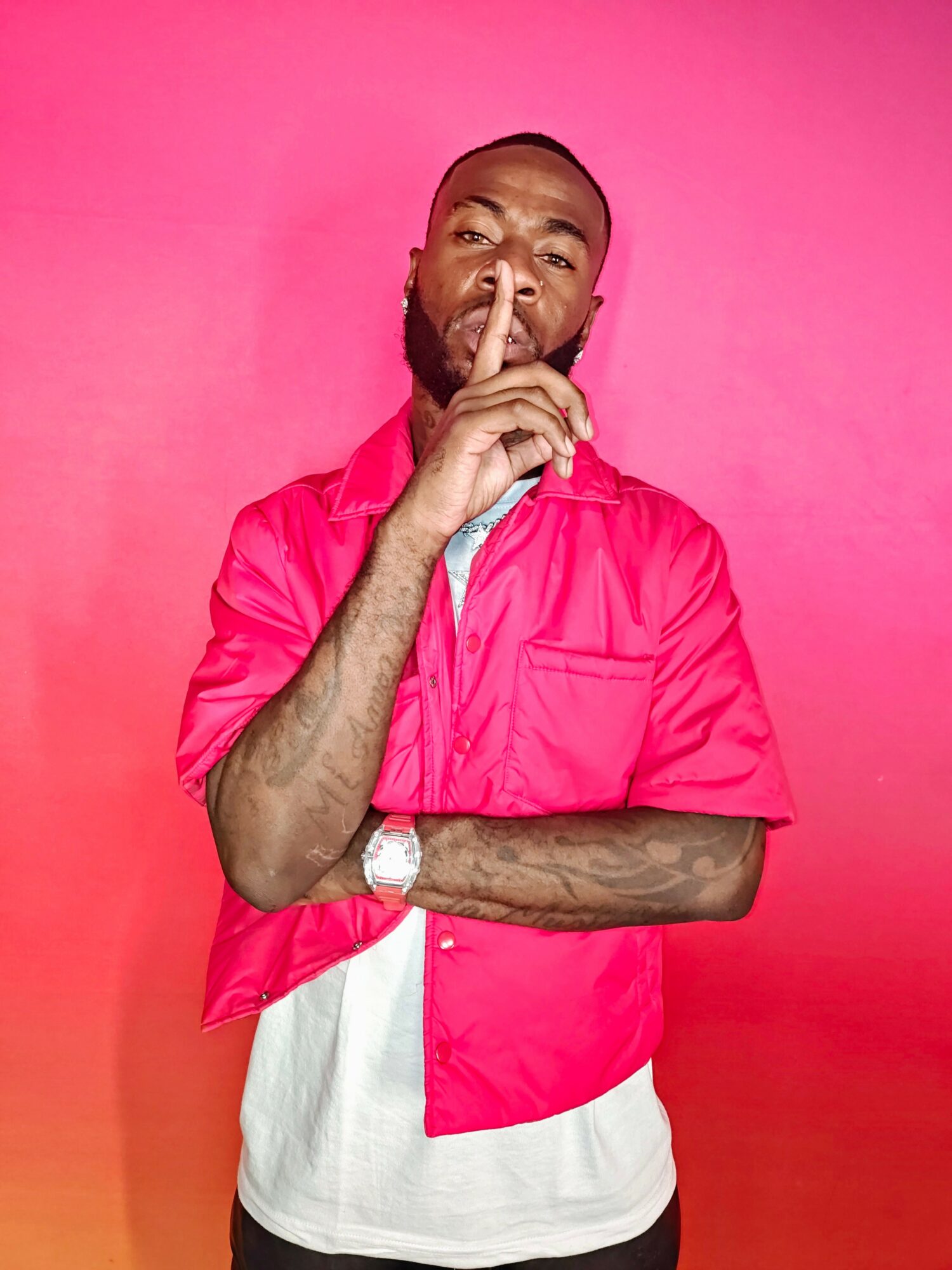 Man with a beard and short hair holding a finger to lips, wearing a pink shirt and white pants, standing against pink background.