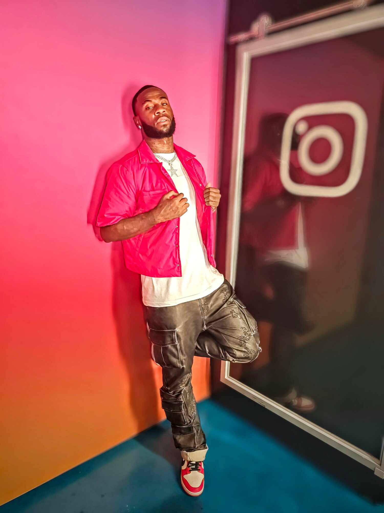 Young man with a beard leaning against a colorful wall, smiling, wearing a pink jacket, white shirt, and jeans.