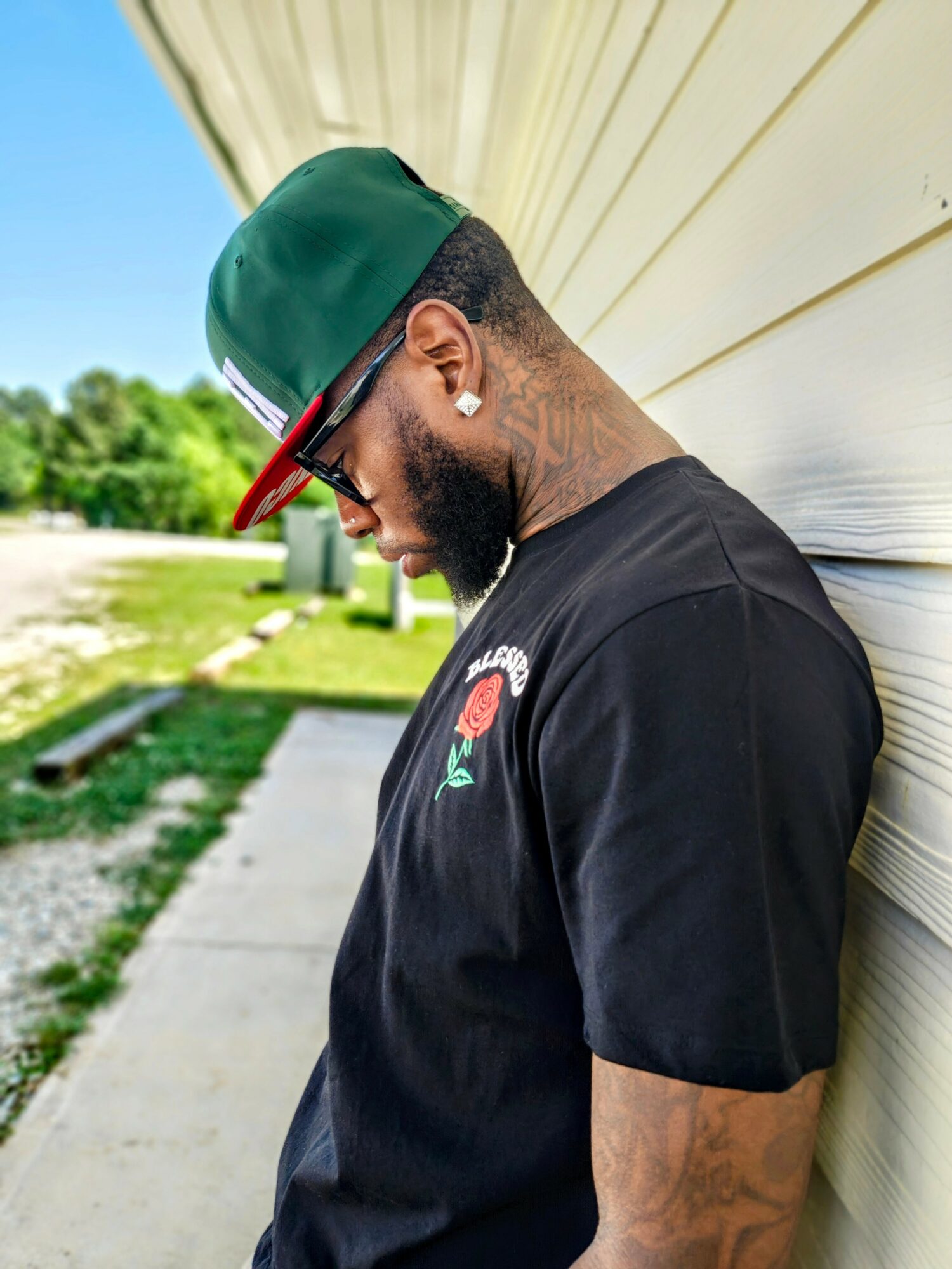 Young man with a beard wearing a green and red cap, sunglasses, and a black T-shirt, leaning against a wall outdoors.