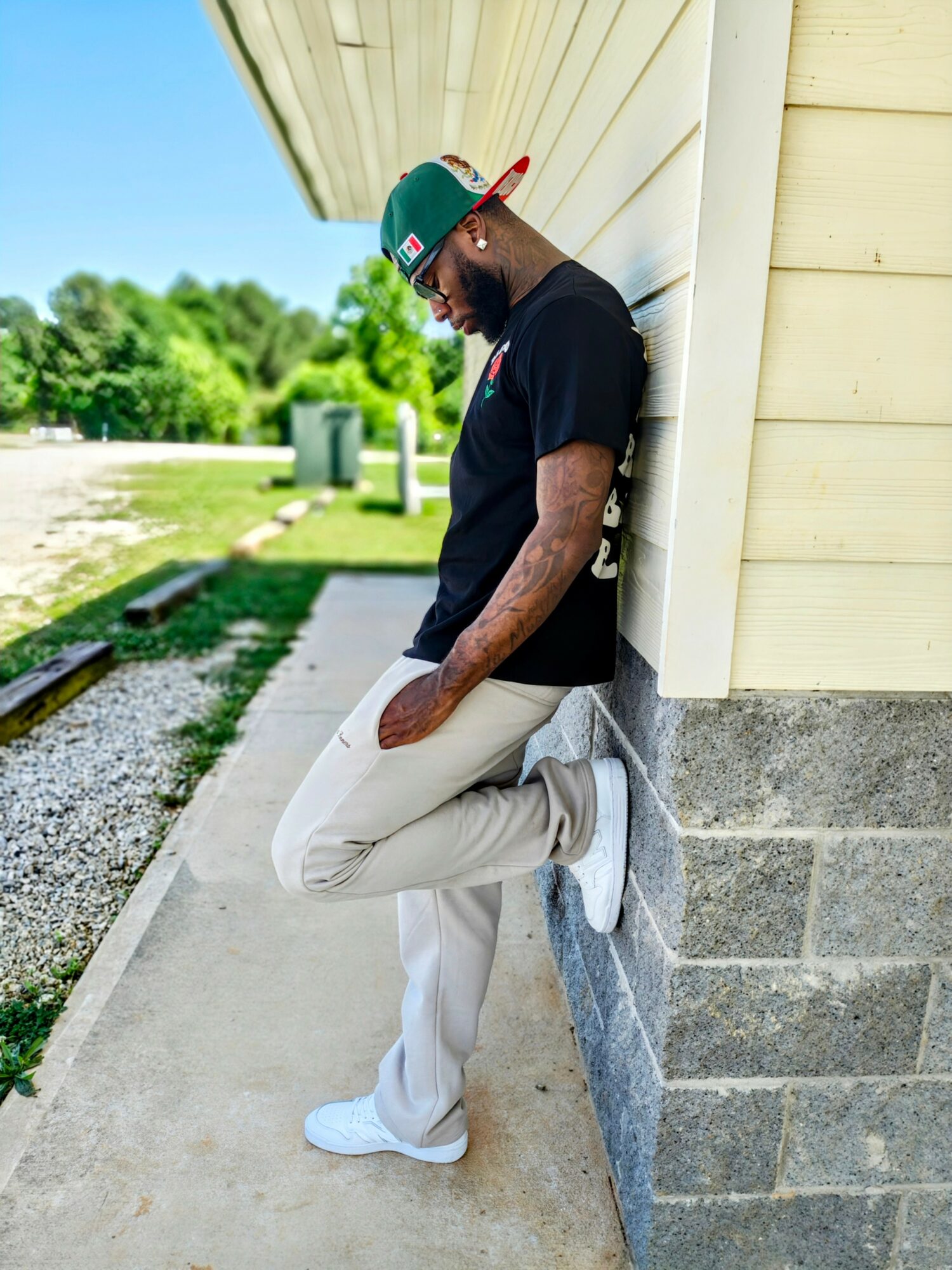 Man leaning against a yellow building outdoors, wearing a cap, sunglasses, black t-shirt, and light-colored pants.