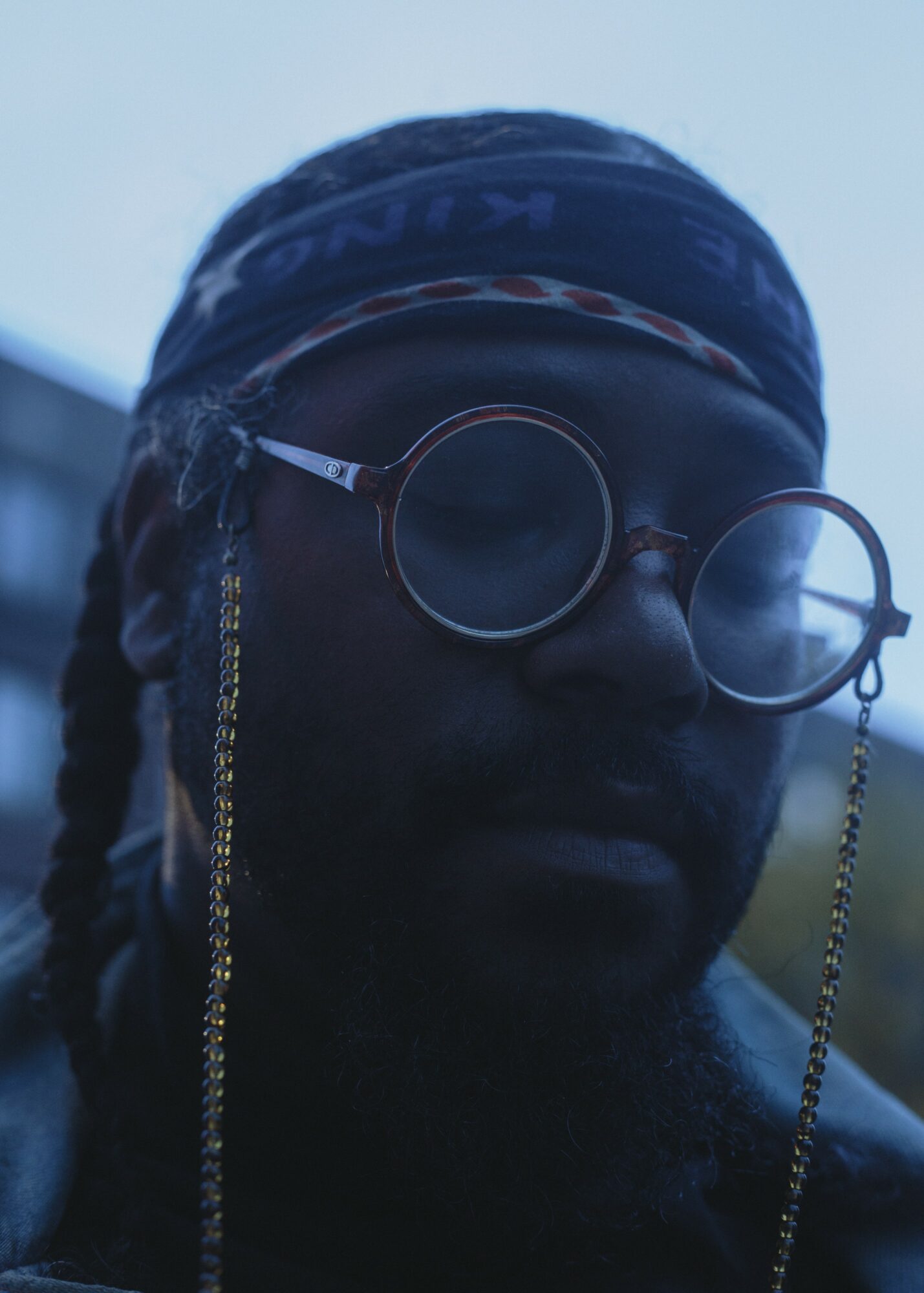 Person wearing round glasses, headscarf, and chain earrings outdoors, with a blurred background.