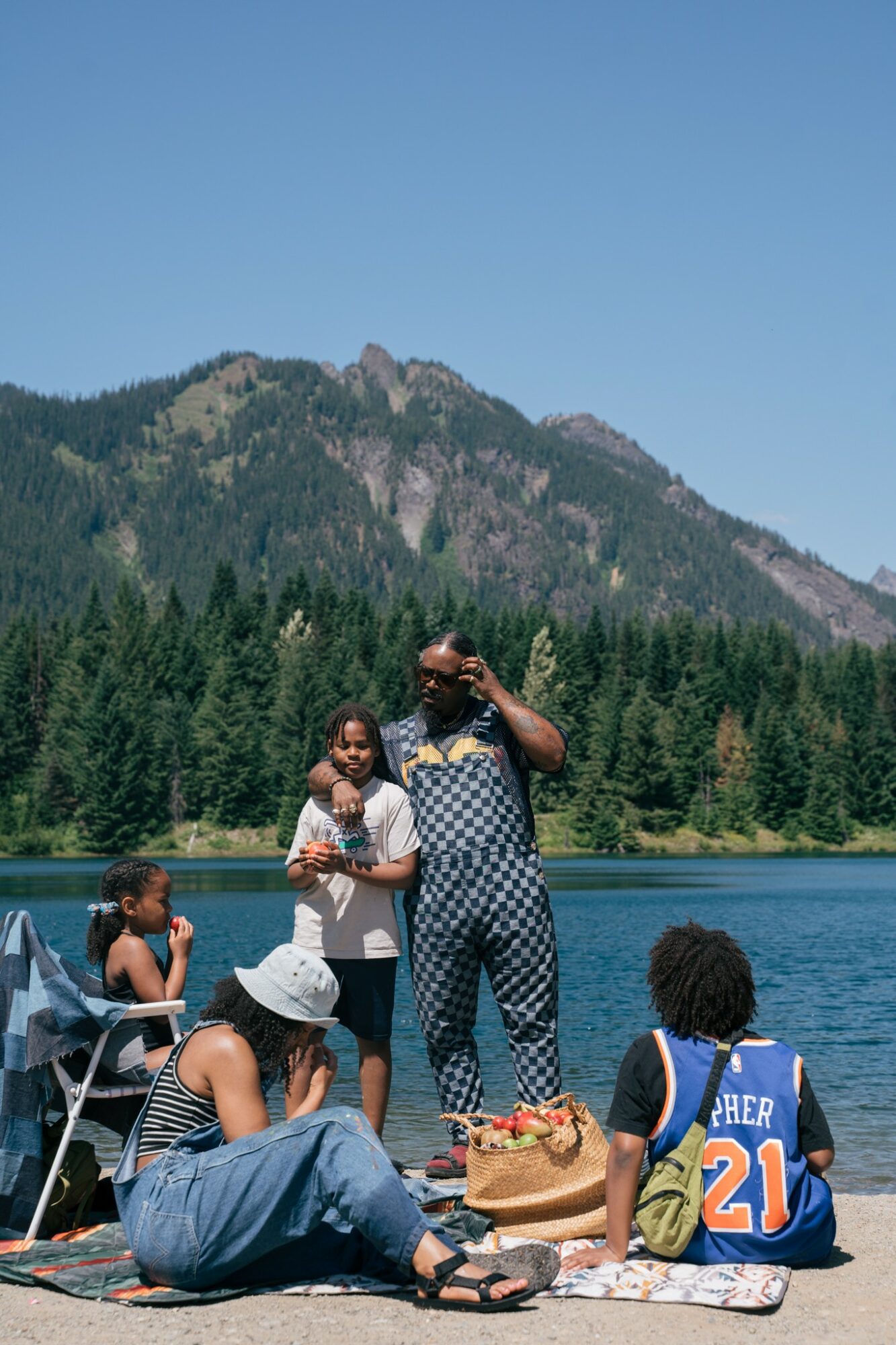 People sitting by a lake with mountains and trees in the background, some standing and others sitting on the ground, enjoying a sunny day.