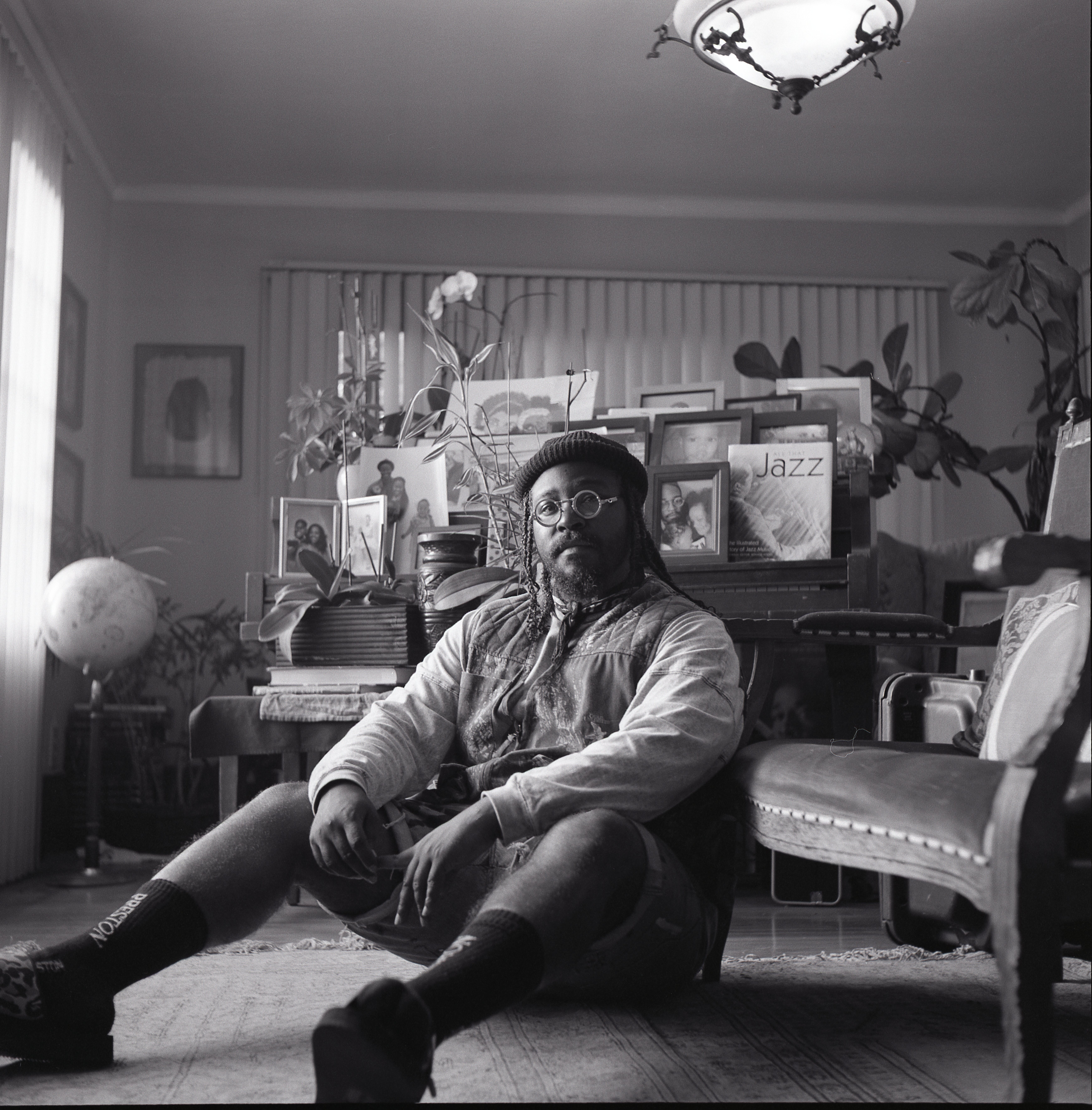 Person with glasses and a hat sitting on the floor in a room filled with plants and framed pictures, black and white photo.