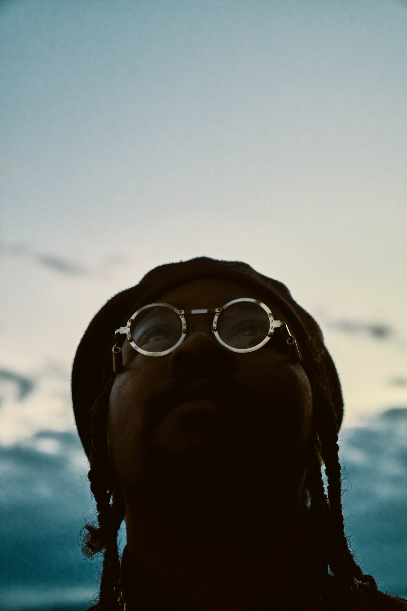 Person with glasses and dreadlocks outdoors, sky and water in background, face mostly in shadow.
