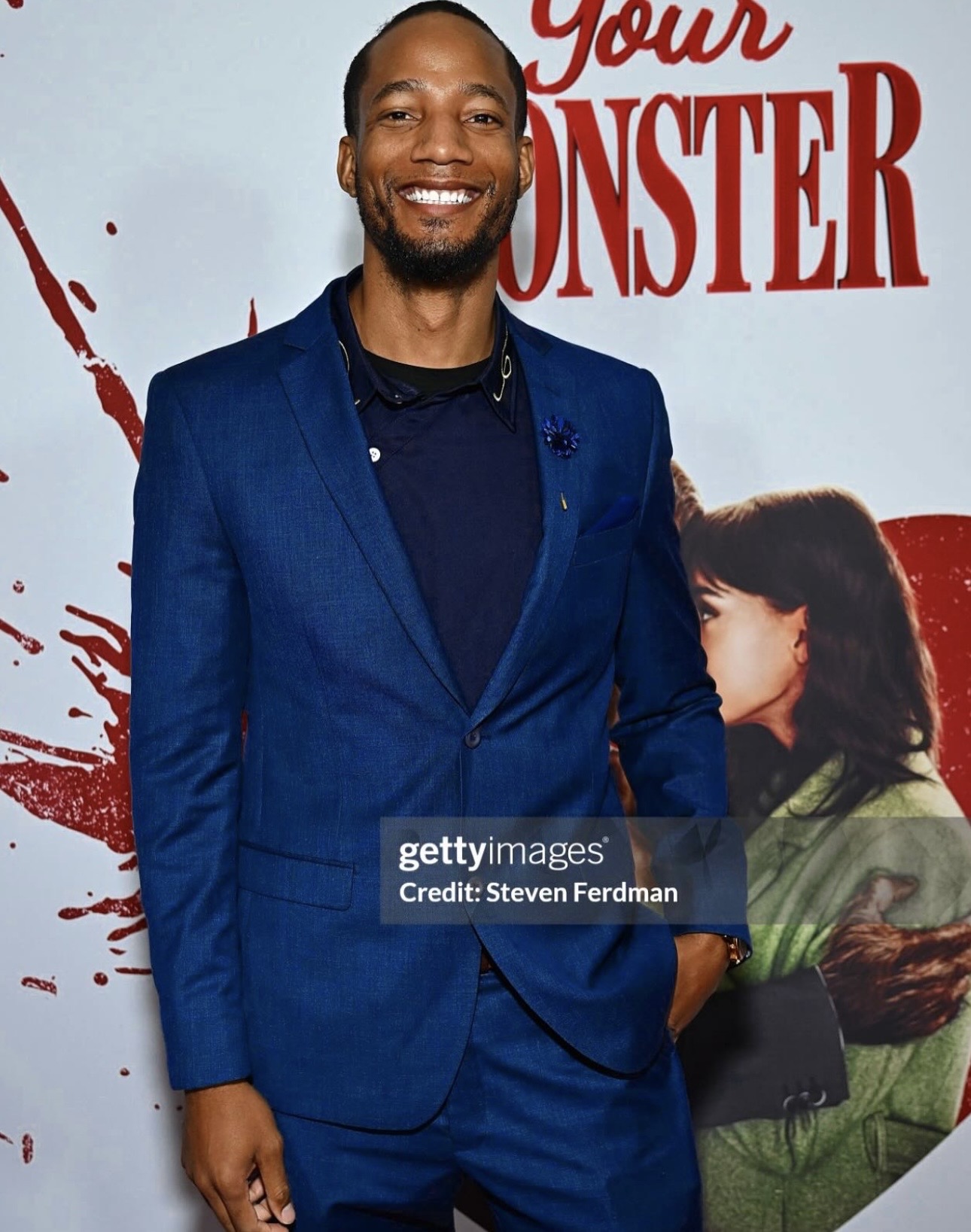 Smiling man in a blue suit standing in front of a movie poster with a girl hugging a monster, red text in background.