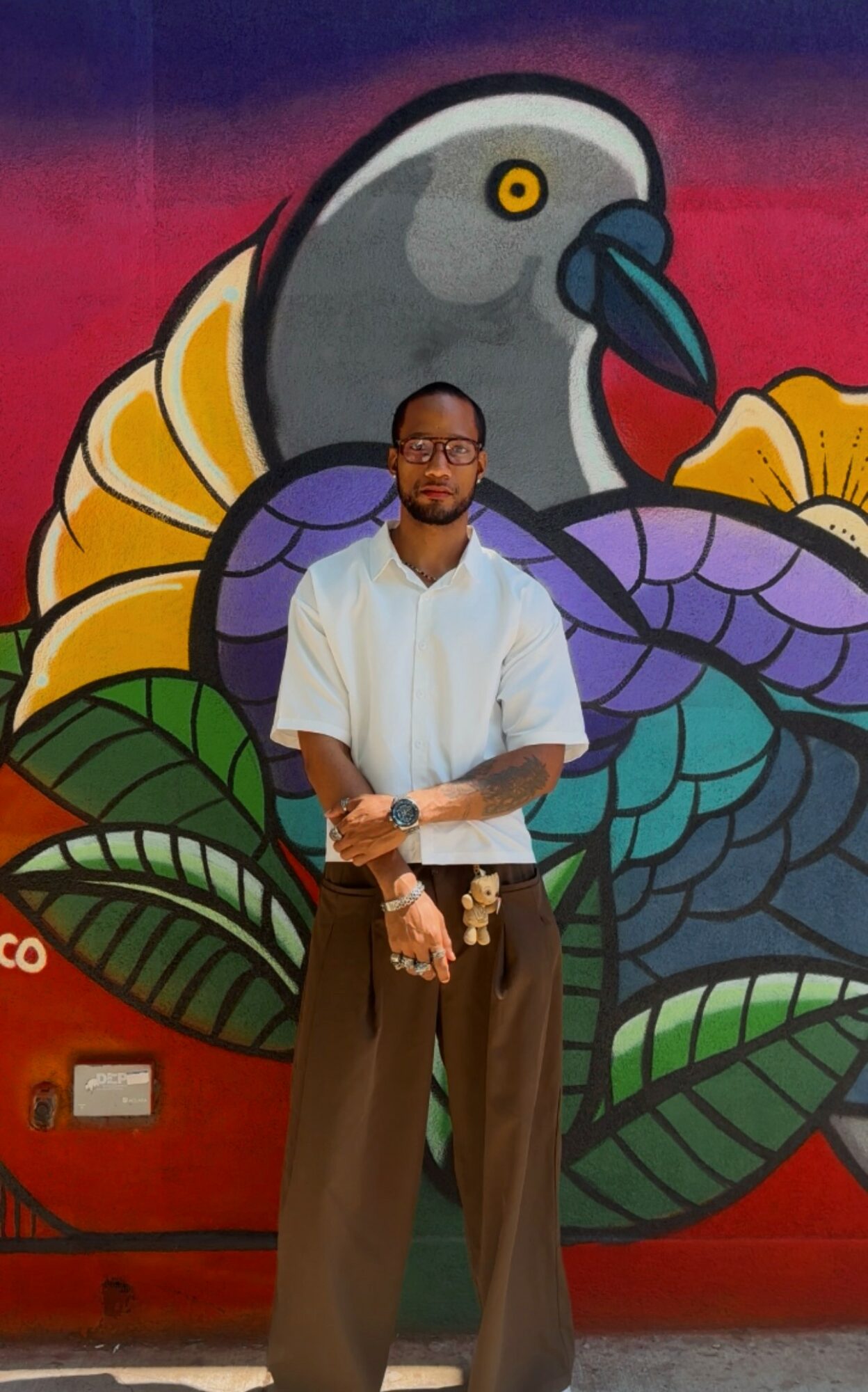 Man with glasses and beard standing in front of colorful mural with a bird and leaves.