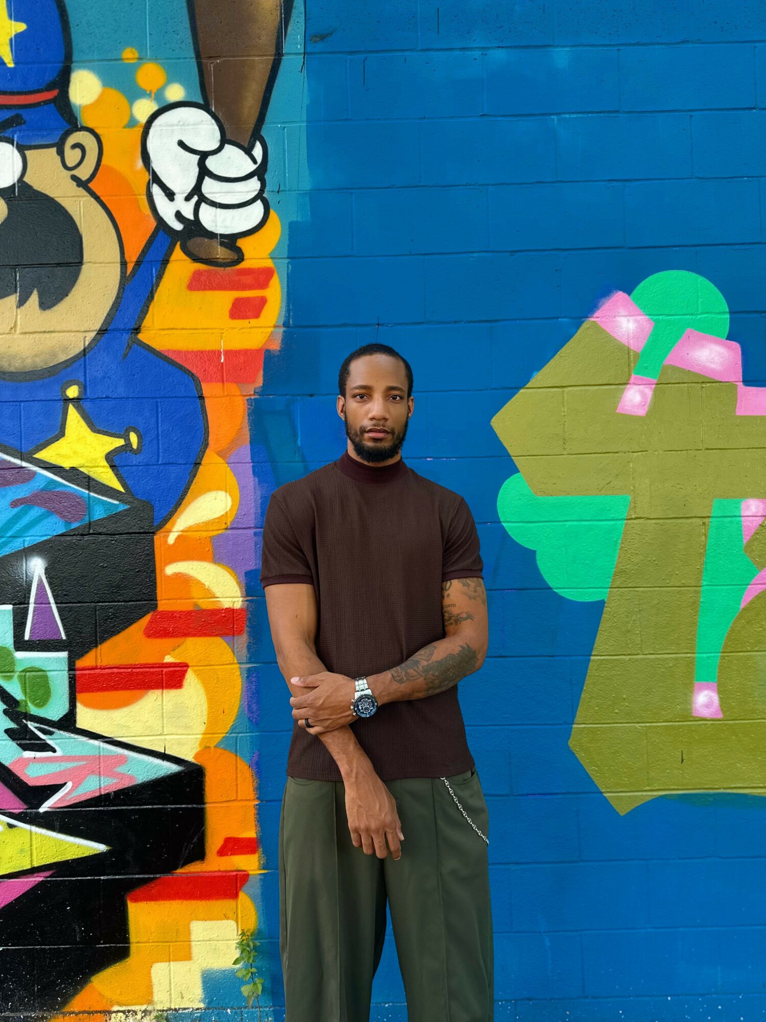 Man with short hair and beard standing in front of colorful graffiti wall, arms crossed, wearing a black t-shirt and gray pants.