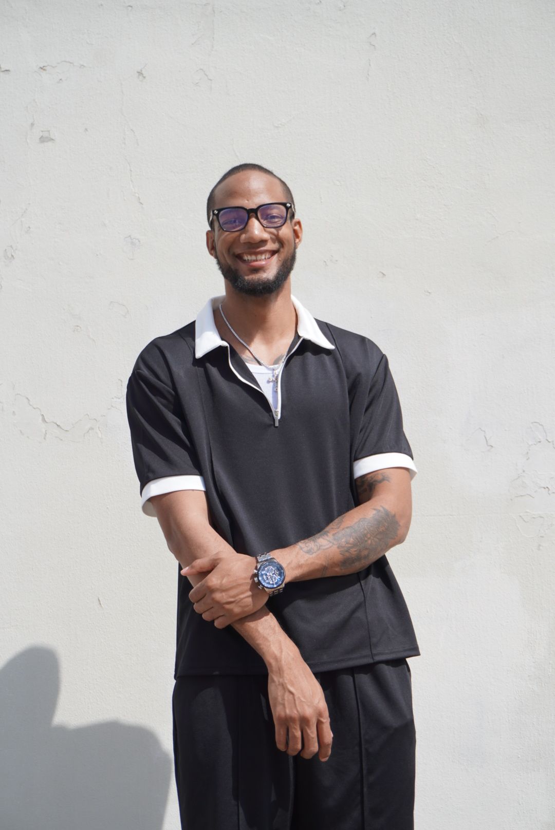 Man with glasses and a watch stands against a plain light background, smiling and looking at the camera.