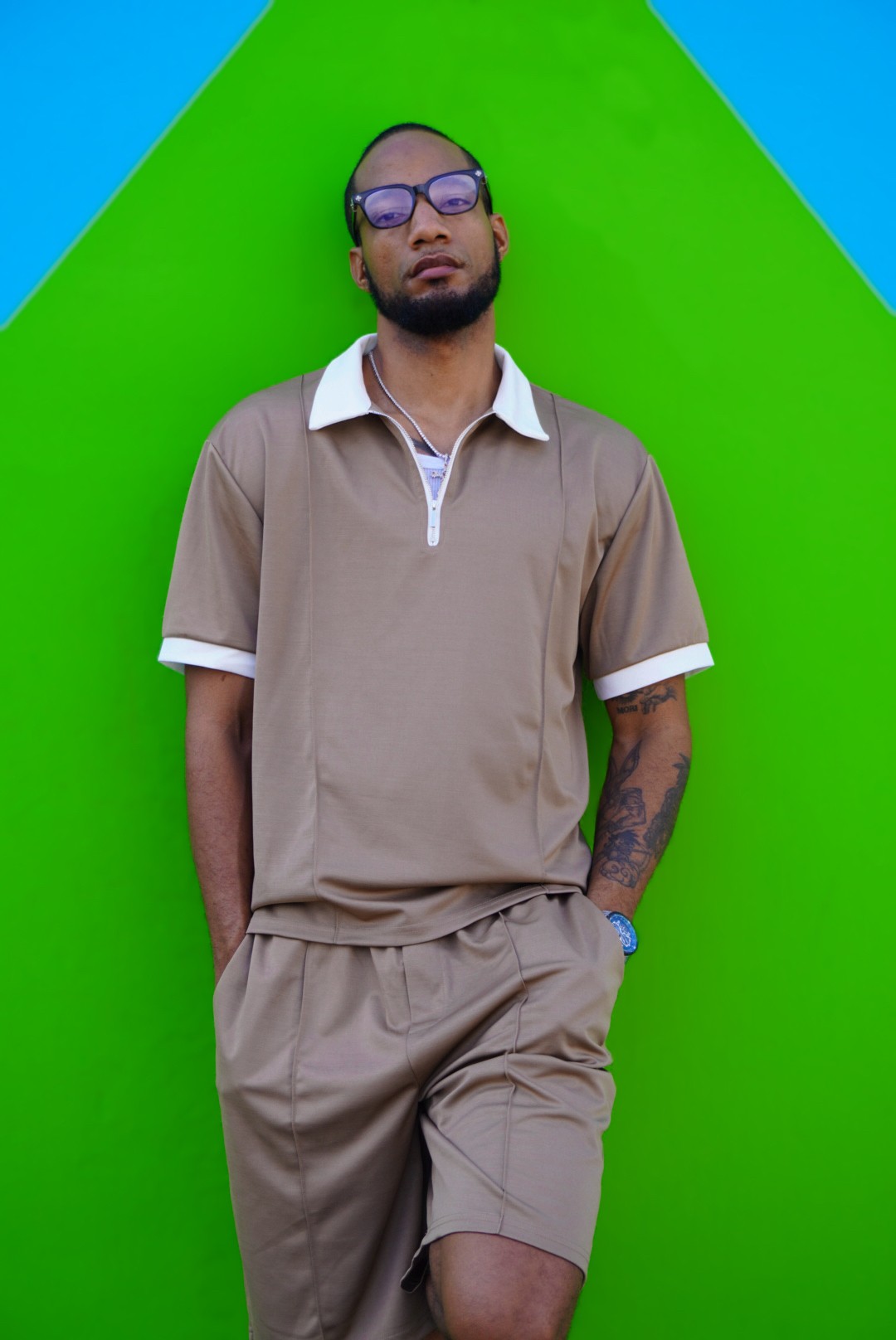 Young man with glasses and a beard stands against a green and blue background, wearing a beige shirt and shorts.