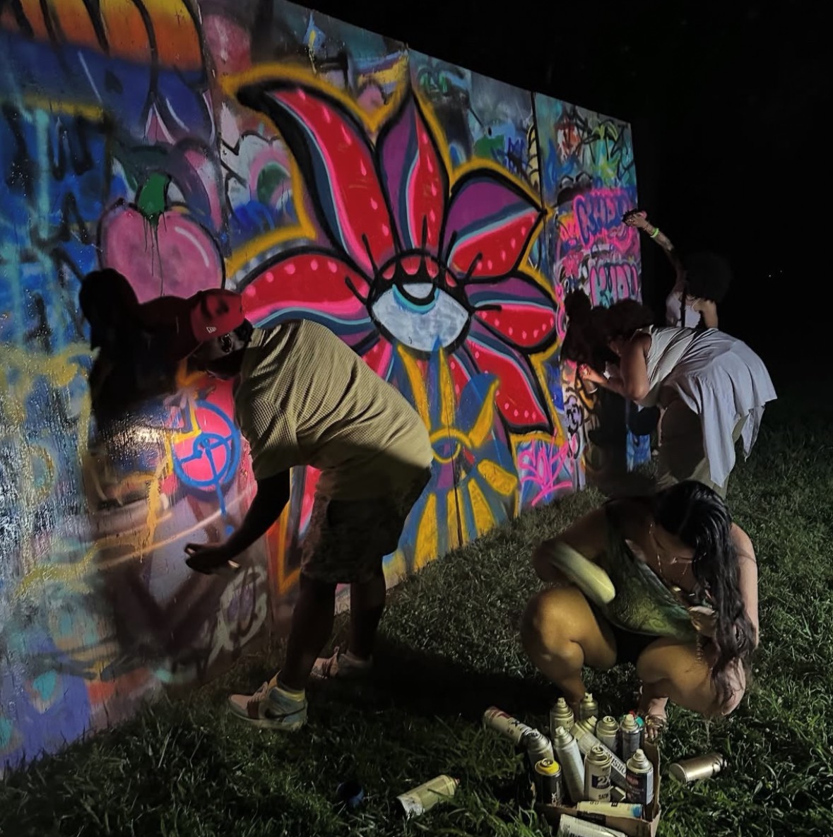 People painting graffiti on a colorful wall at night, with spray cans on the ground, and a grassy area nearby.
