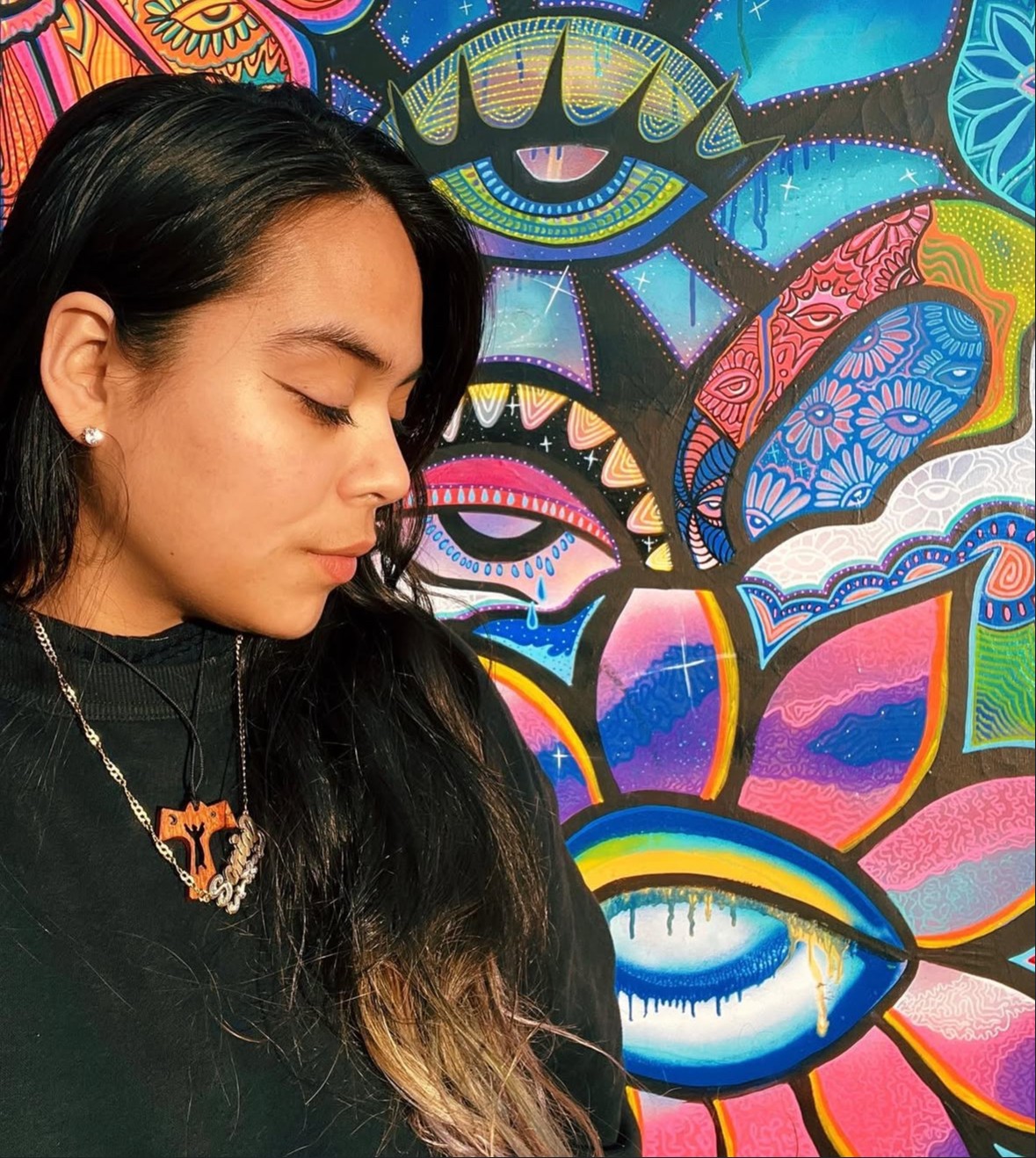 Young woman with long dark hair and earrings, wearing a black top and necklace, stands in front of colorful abstract mural.