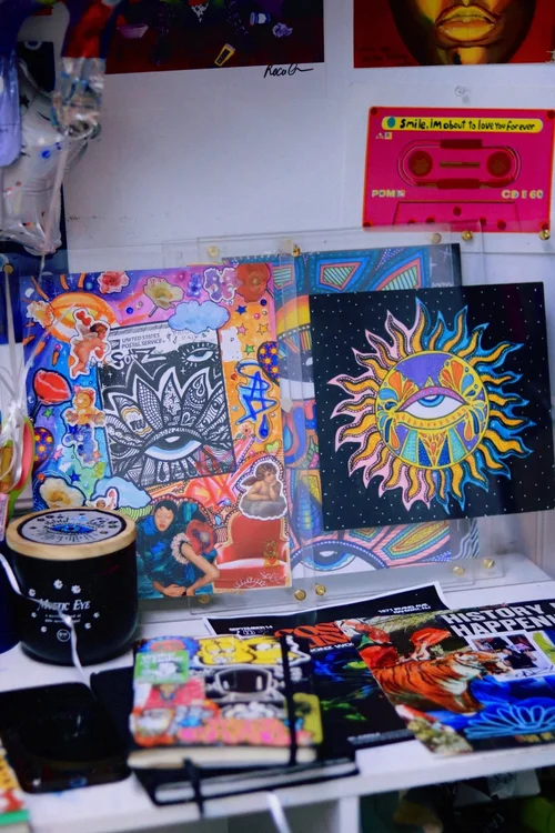 Colorful artwork featuring a sun with a face, displayed on a wall with other art pieces and a pink cassette tape above.