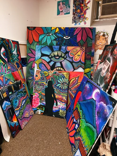 Art display with colorful paintings leaning against a wall, including abstract and nature-inspired designs, with a person viewing artwork.