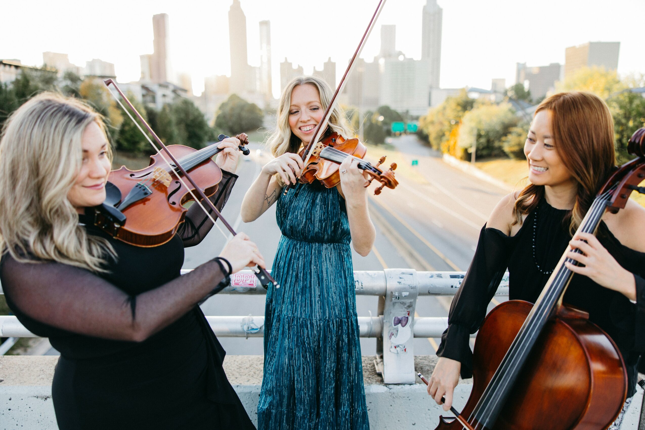 Three women playing violins and a cello outdoors on a bridge with city skyline in background.