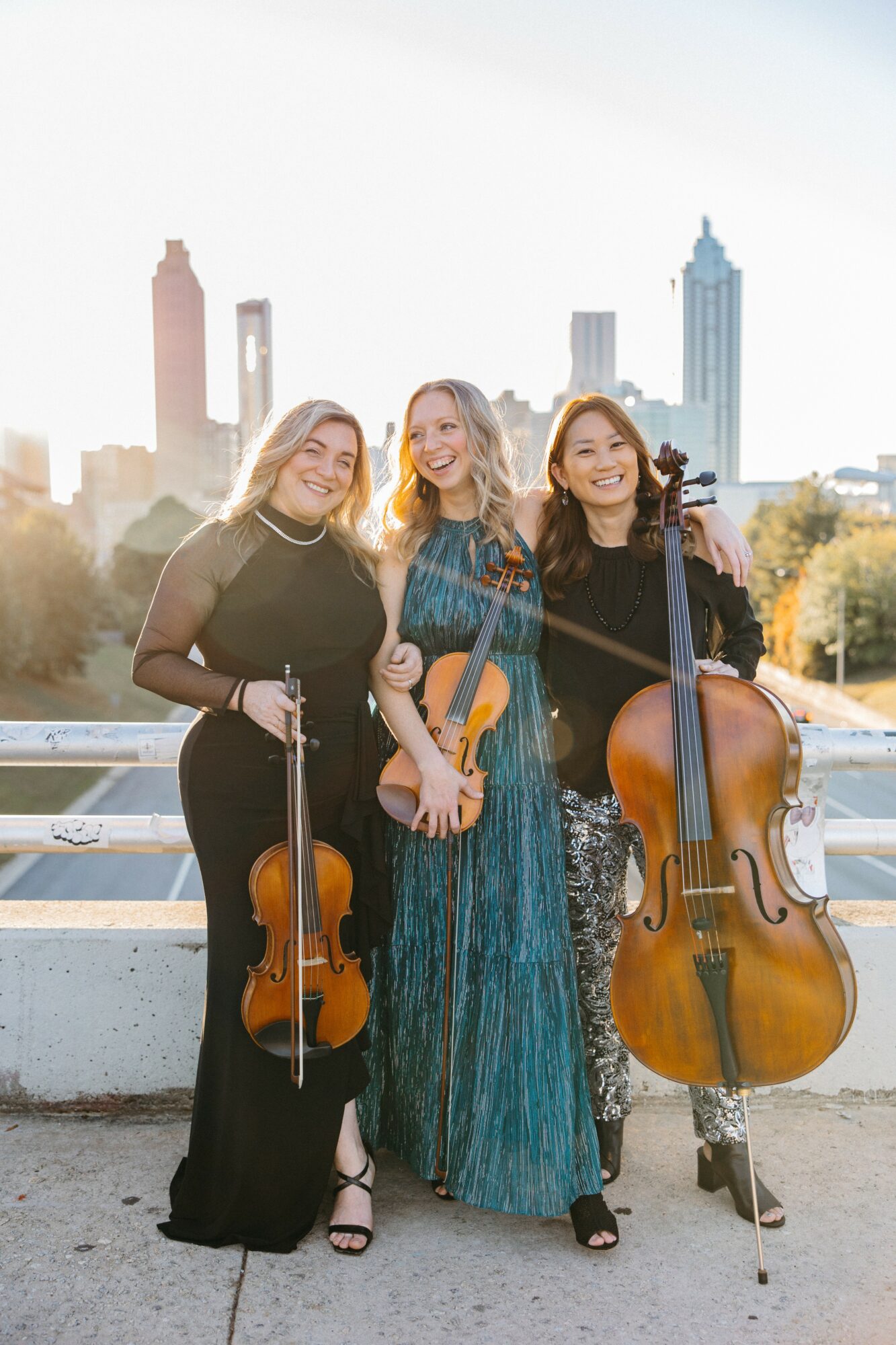 Three women holding string instruments, standing outdoors with city skyline in background, sunlight shining behind them.