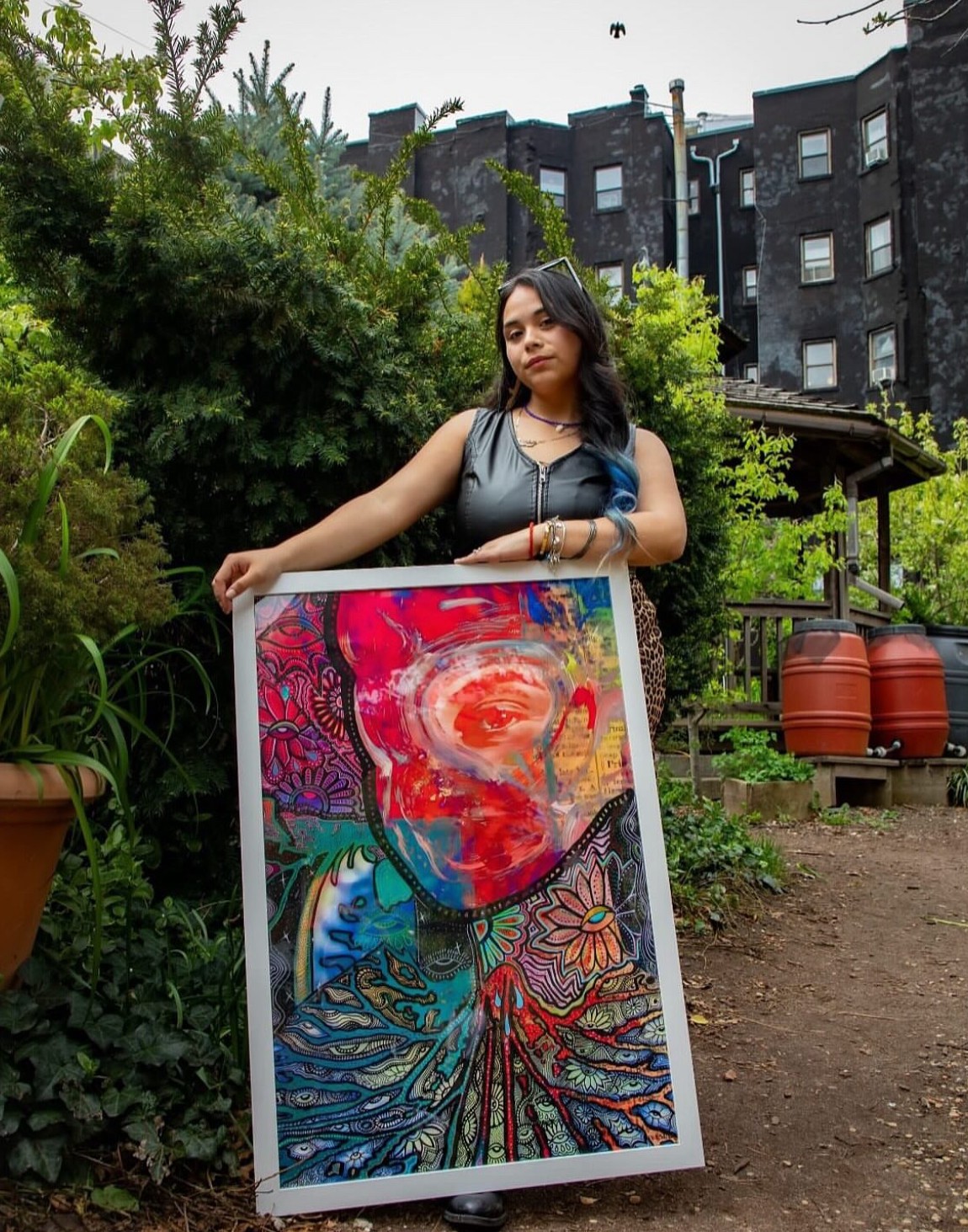 Young woman standing outdoors holding a colorful abstract painting, surrounded by greenery and buildings in the background.