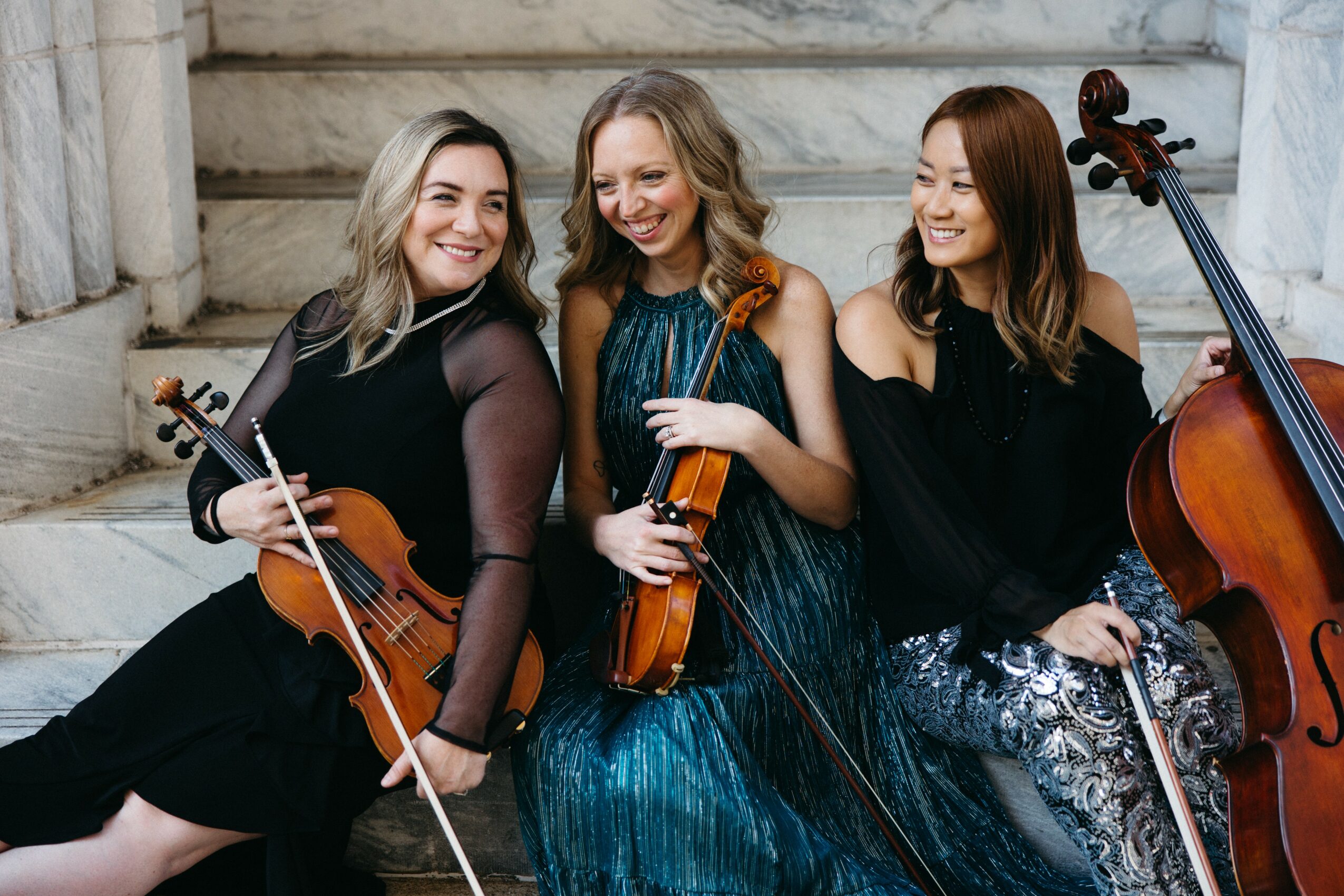 Three women holding violins and a cello, sitting on stone steps, smiling and looking at each other.