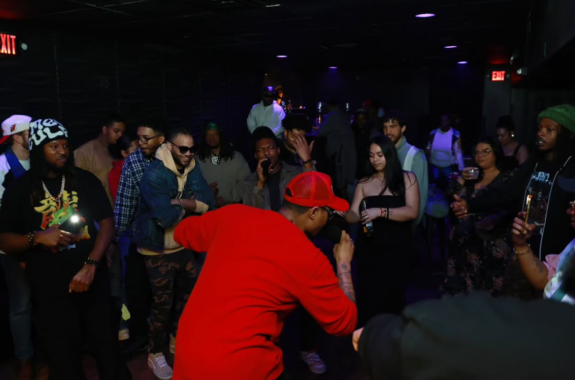 Group of people dancing and socializing in a dimly lit nightclub with purple lighting, some holding drinks, diverse ages and styles.