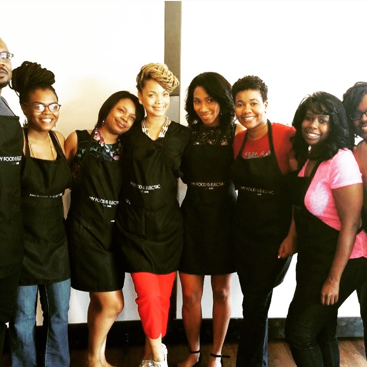 Group of seven women standing together, smiling, wearing aprons, in a bright room with a white wall.
