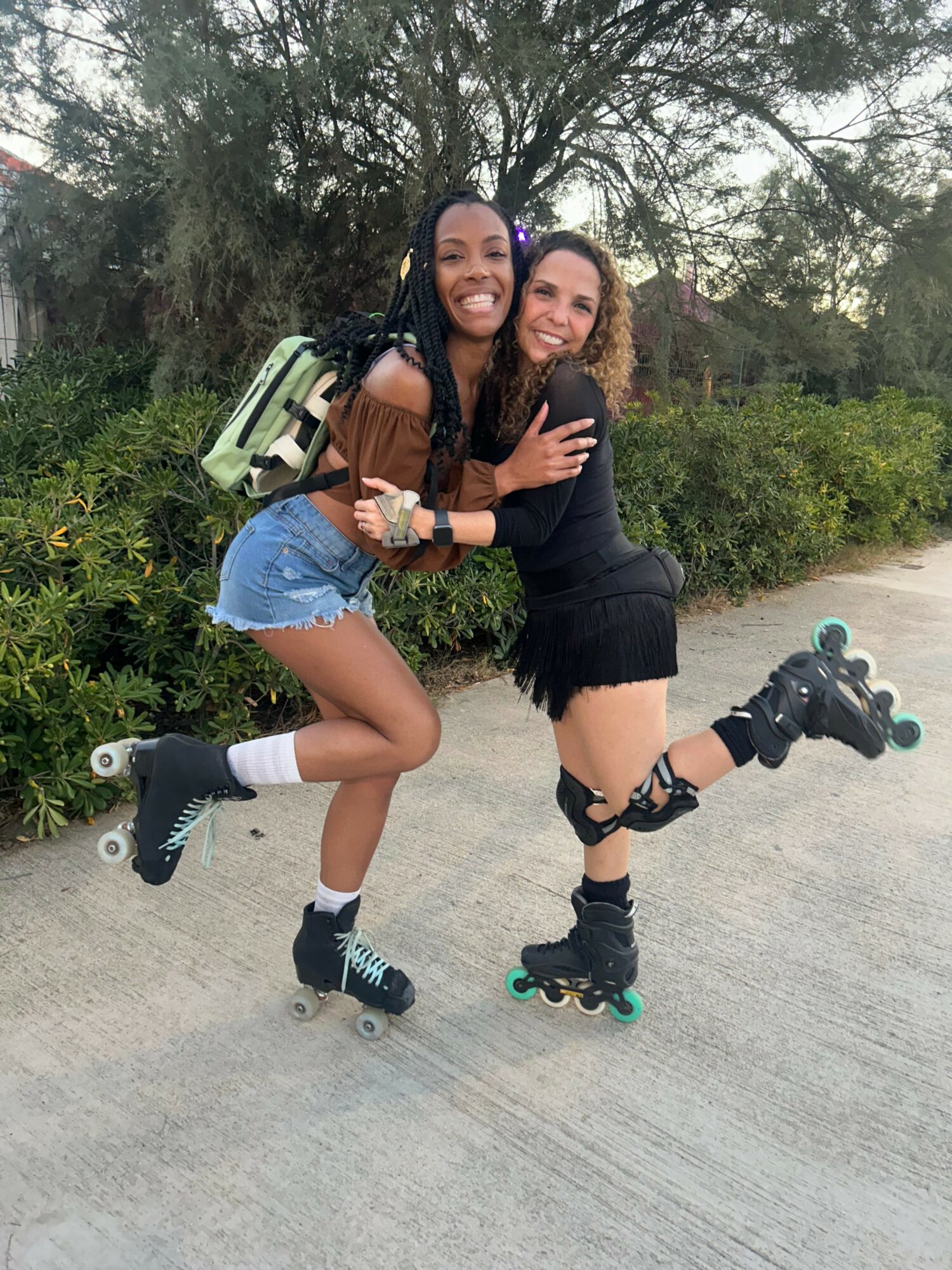 Two women wearing roller skates, smiling and hugging outdoors on a concrete path with trees and bushes in background.