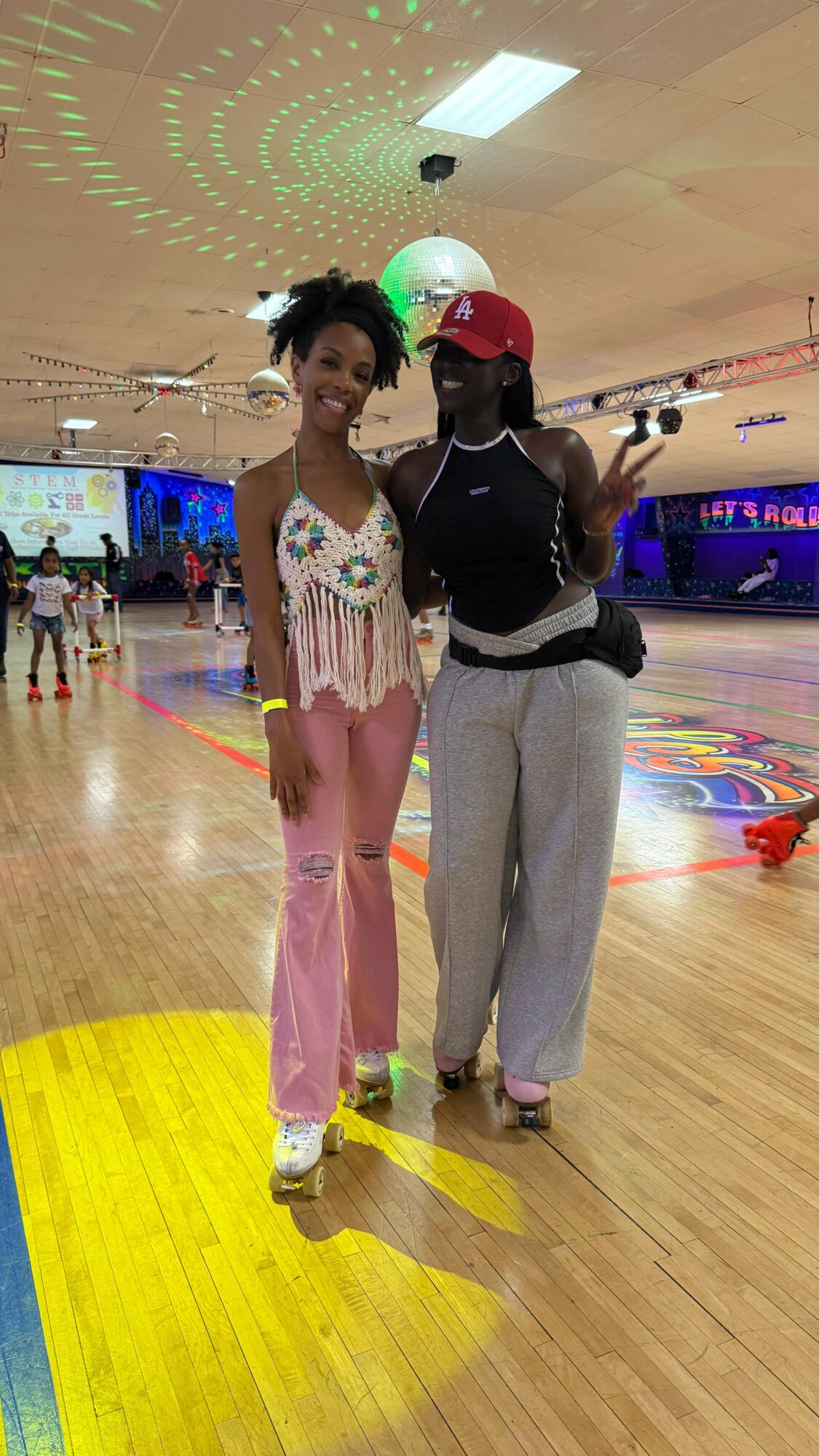 Two women stand together in a roller skating rink, smiling, with colorful lights and a screen in the background.