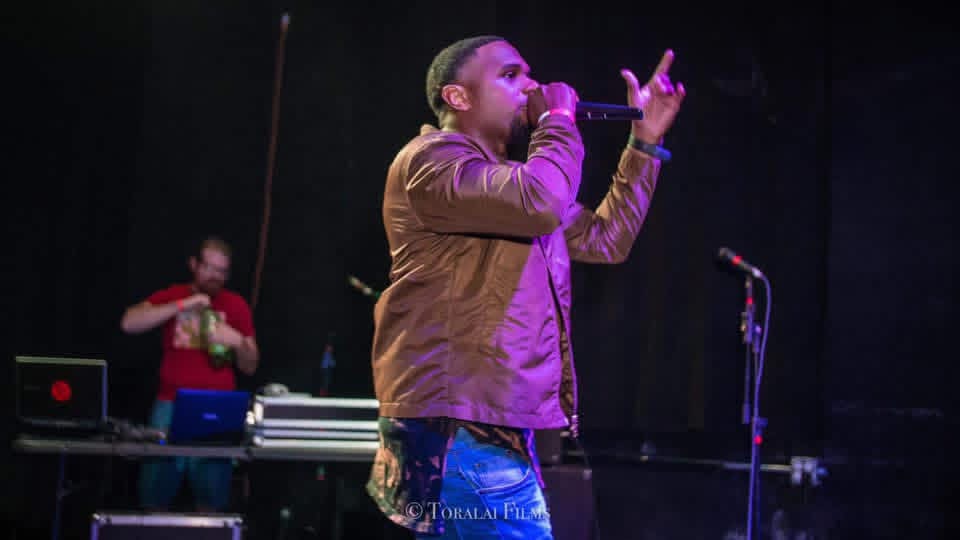 Male singer performs on stage with a microphone, with a DJ in the background, dark stage lighting.
