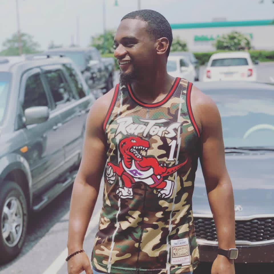 Smiling man in camouflage tank top with a cartoon dinosaur, standing in parking lot with cars in background.
