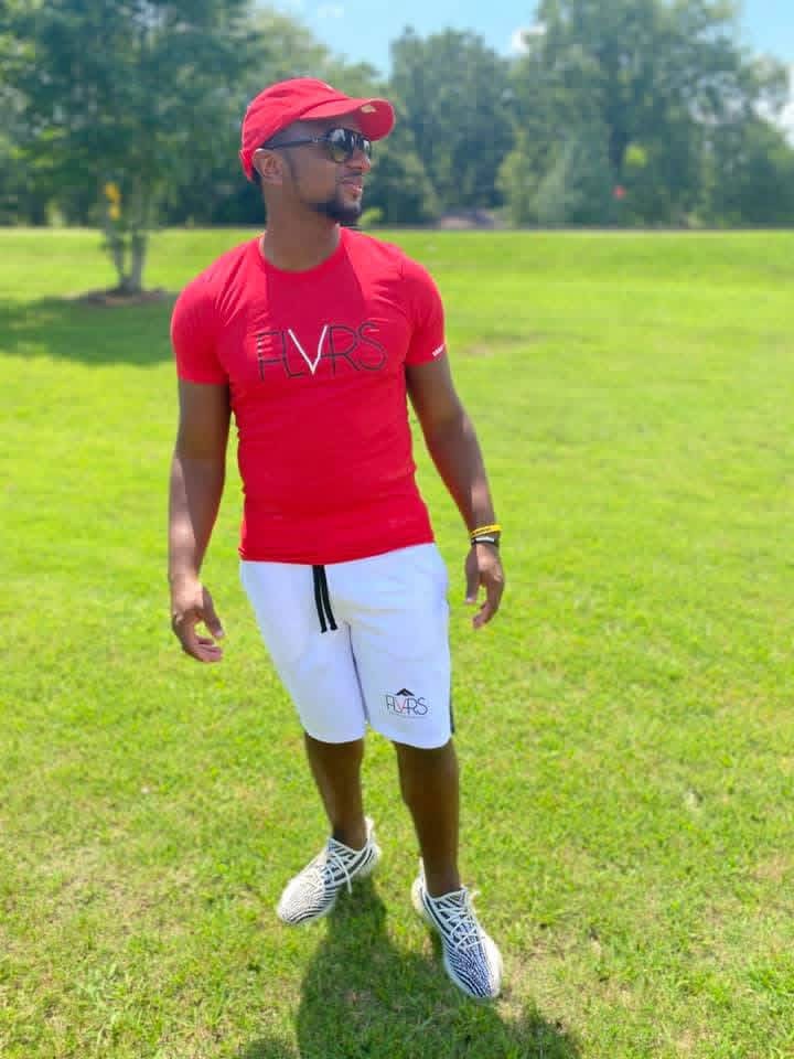Man in red cap, sunglasses, red t-shirt, white shorts, and sneakers walking on grass in park.