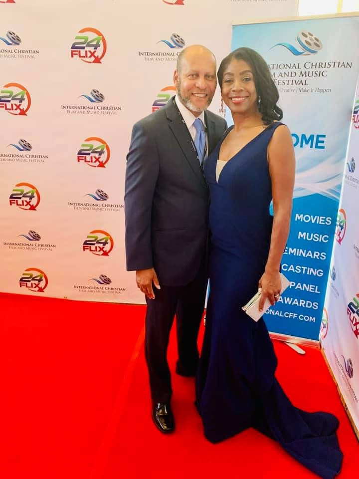 Man and woman in formal attire standing on red carpet, smiling, with event banners in background.