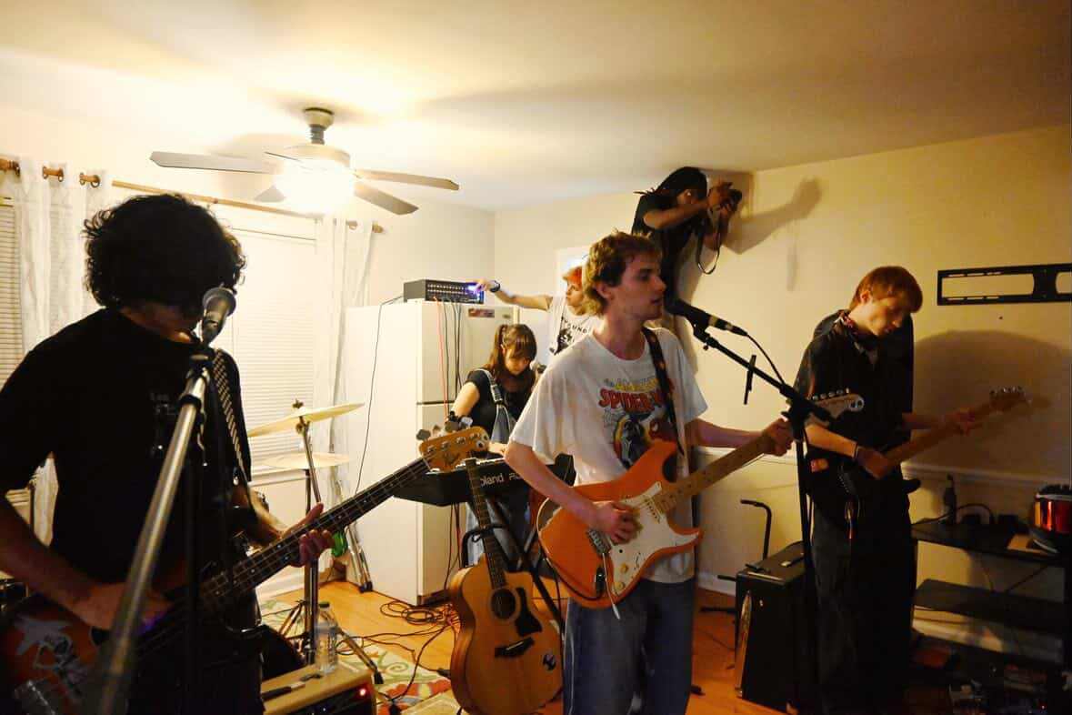 Group of five people playing musical instruments in a room, with one person on a ladder adjusting something on the wall.