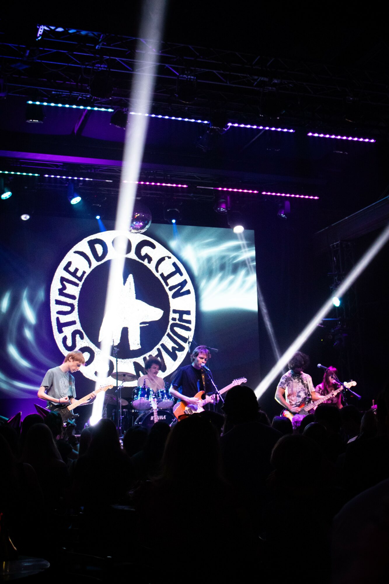 Band performs on stage with a large circular logo in the background, colorful lights, and audience silhouettes visible.