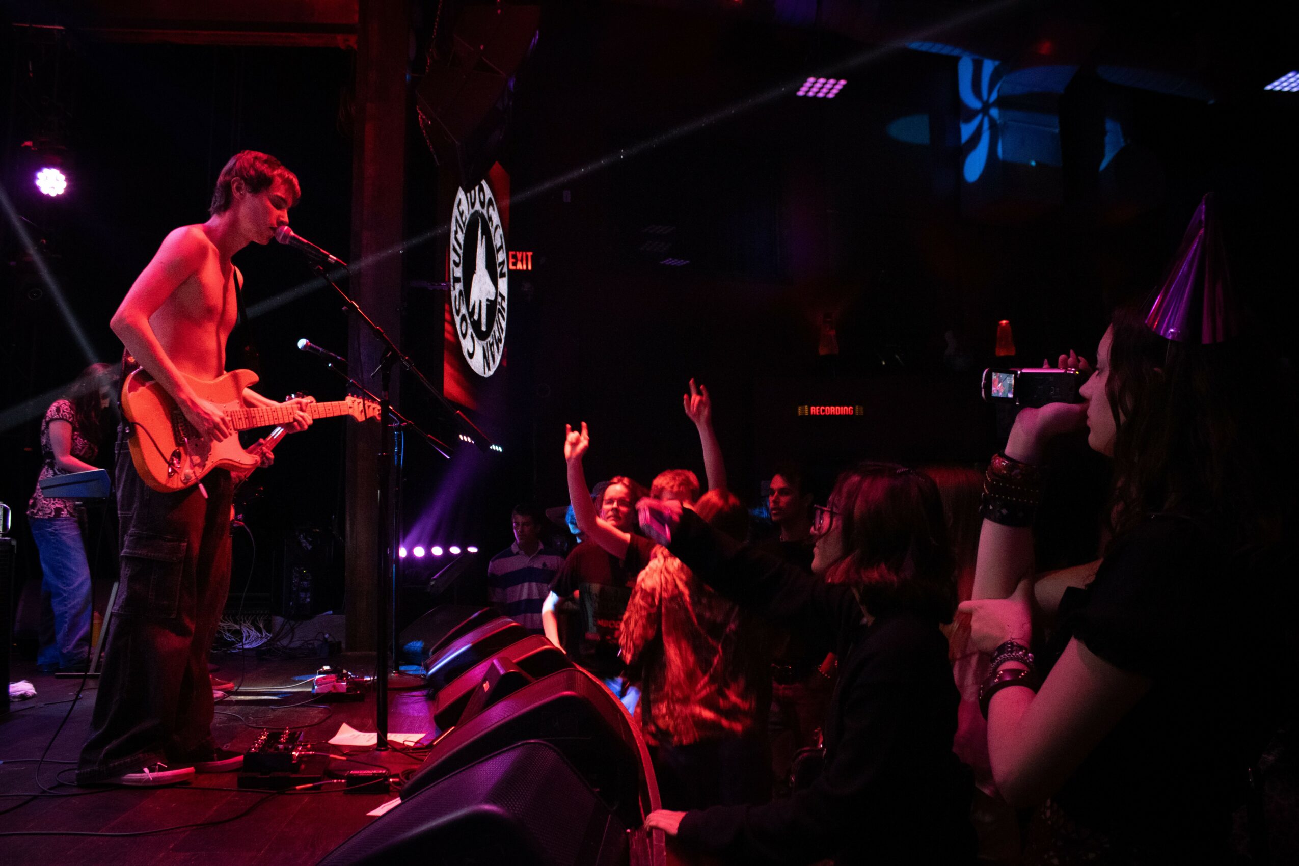 Musician playing guitar on stage with audience raising hands, dark background with stage lights, and a screen displaying a logo.