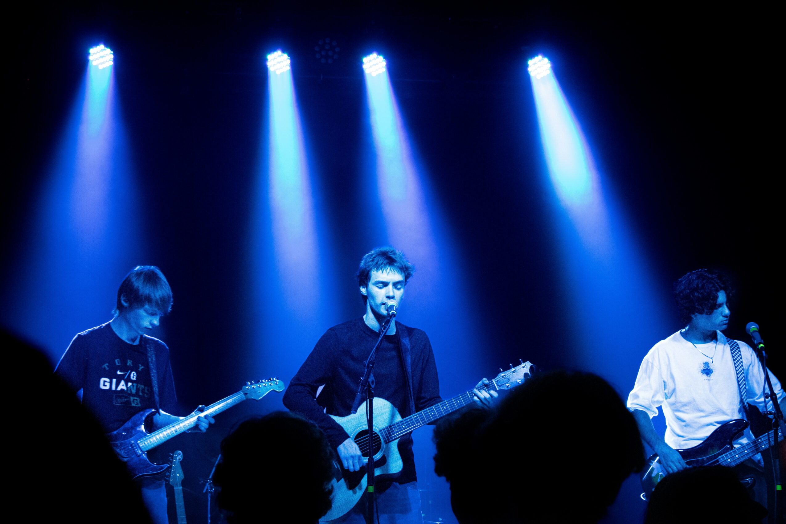 Three musicians perform on stage under blue spotlights, with silhouettes of audience members in foreground.