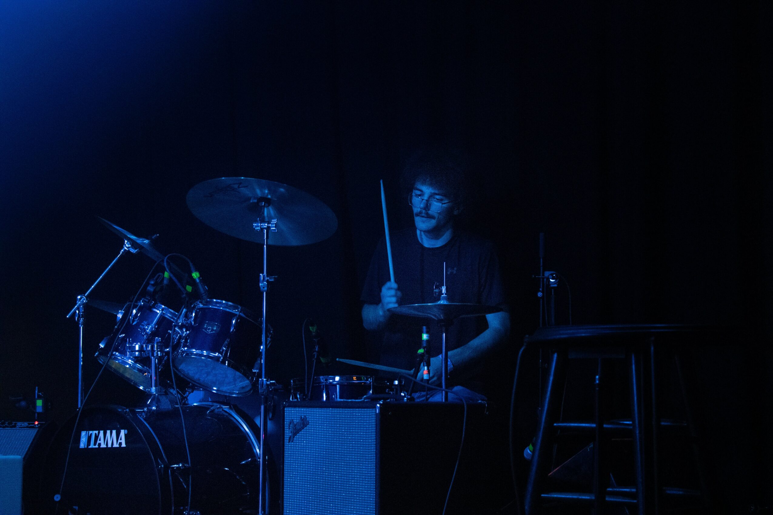 Person playing drums on a dark stage with blue lighting, surrounded by musical equipment.